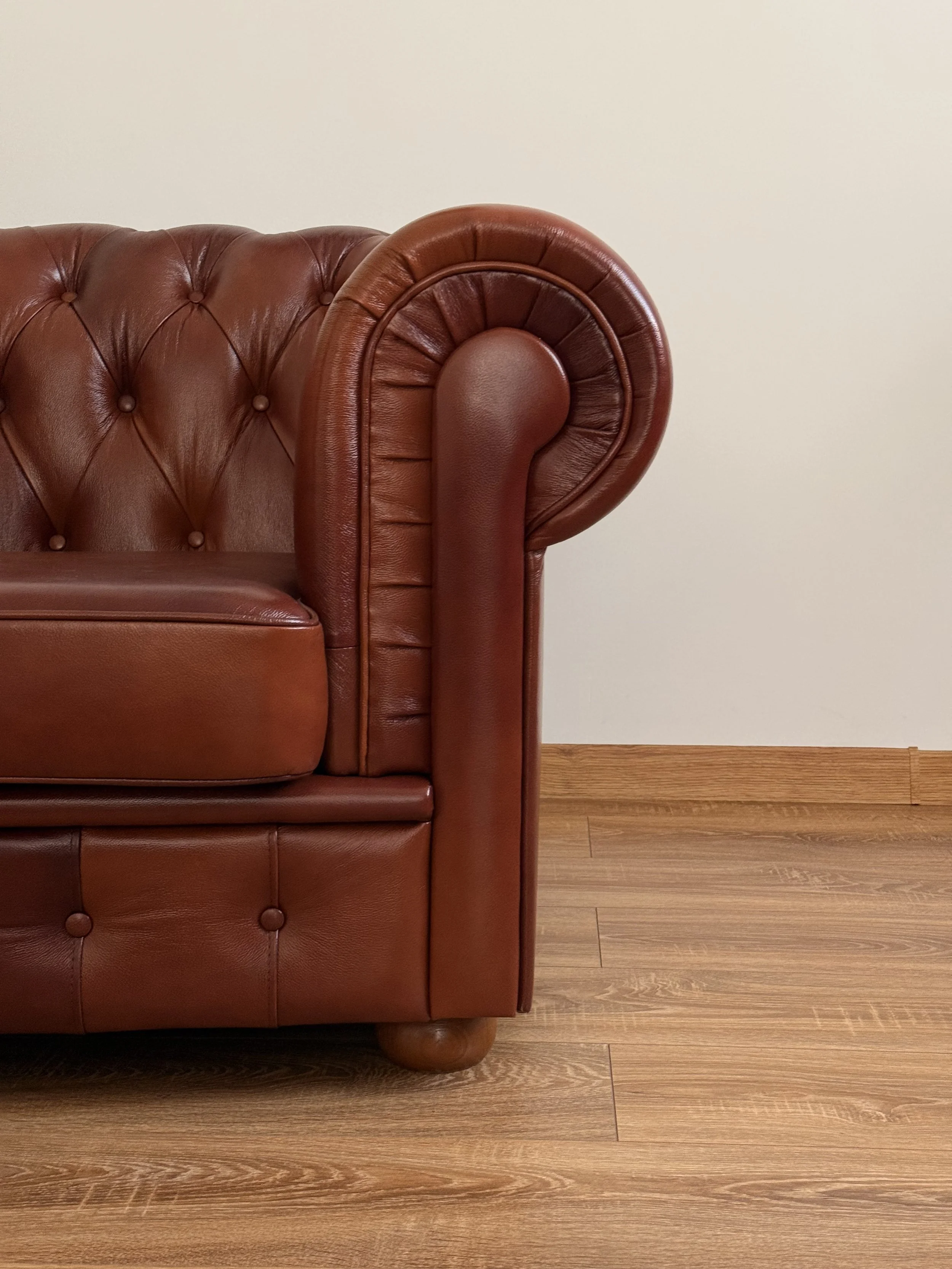 Close-up of a brown leather tufted sofa against a plain beige wall with wooden flooring.