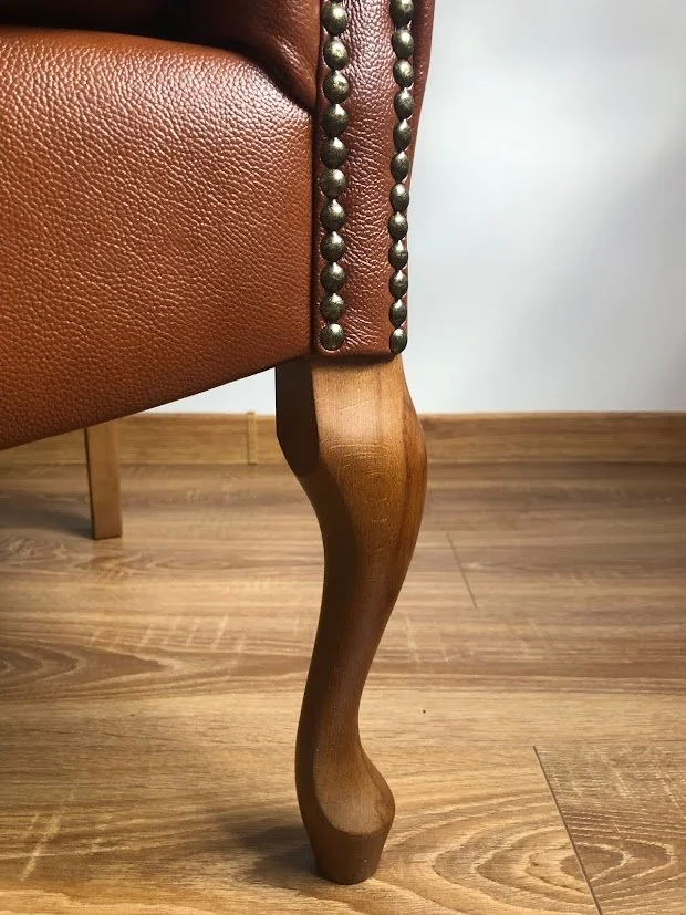 Close-up of a vintage-style armchair with a wooden cabriole leg, brown leather upholstery, and decorative nailhead trim.