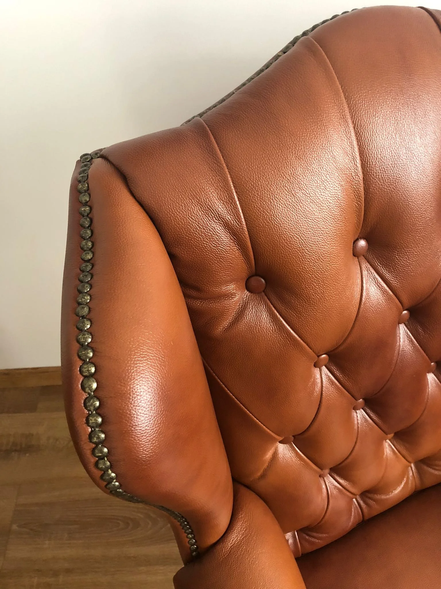 Close-up of a brown leather tufted armchair with nailhead trim against a plain wall and wooden floor.