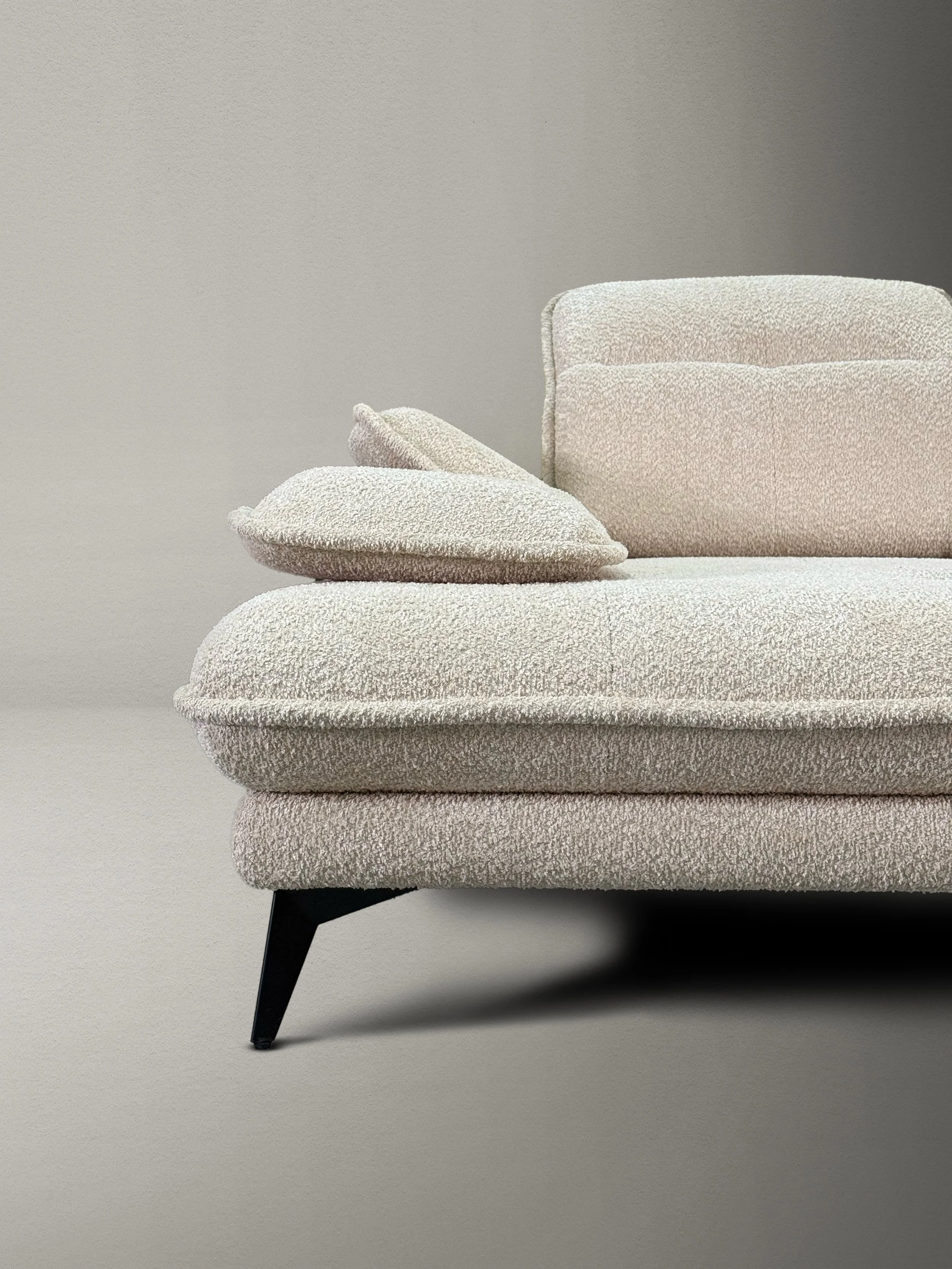 Close-up of a modern beige upholstered sofa with a matching cushion, black angled leg, against a plain, light-colored wall.