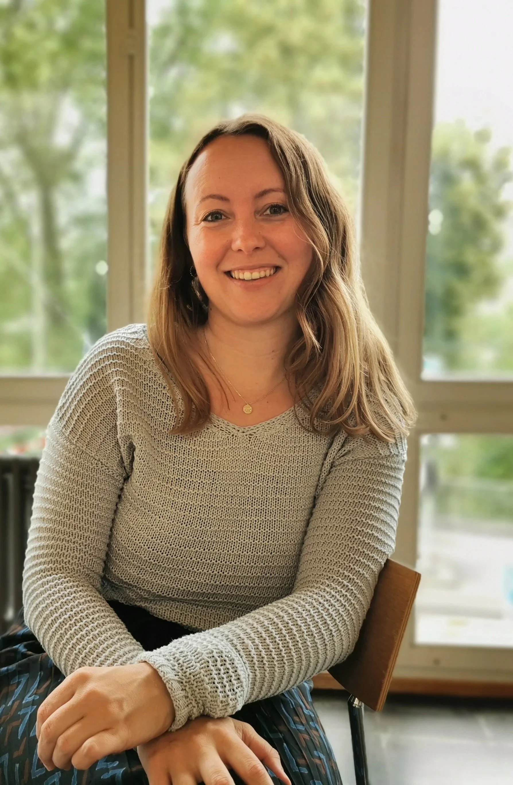 Eine lächelnde Frau mit braunen, gewellten Haaren sitzt in einem Raum mit großen Fenstern, durch die grüne Bäume sichtbar sind.