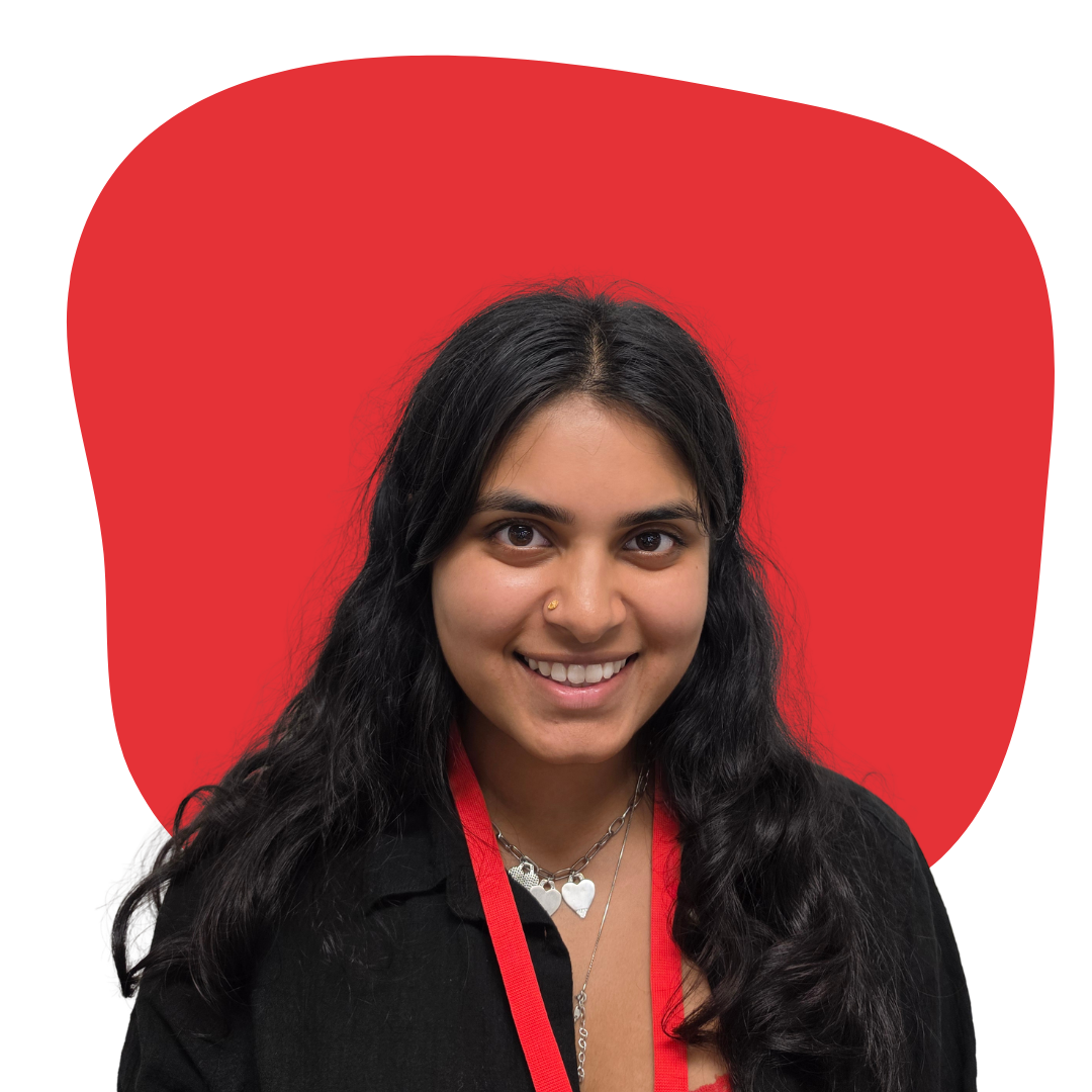 A young woman with long dark hair and light skin, smiling, wearing a necklace with heart-shaped pendants, a black top, and a red lanyard, against a red background.