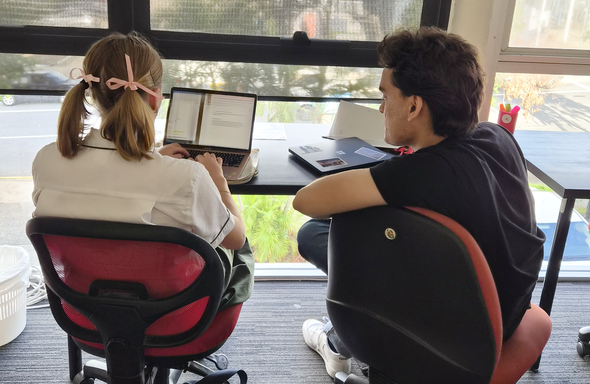 A young girl with blonde hair, tied with pink ribbons, working on a laptop at a desk, with an older man in a black t-shirt observing her, in a room with large windows and natural light.