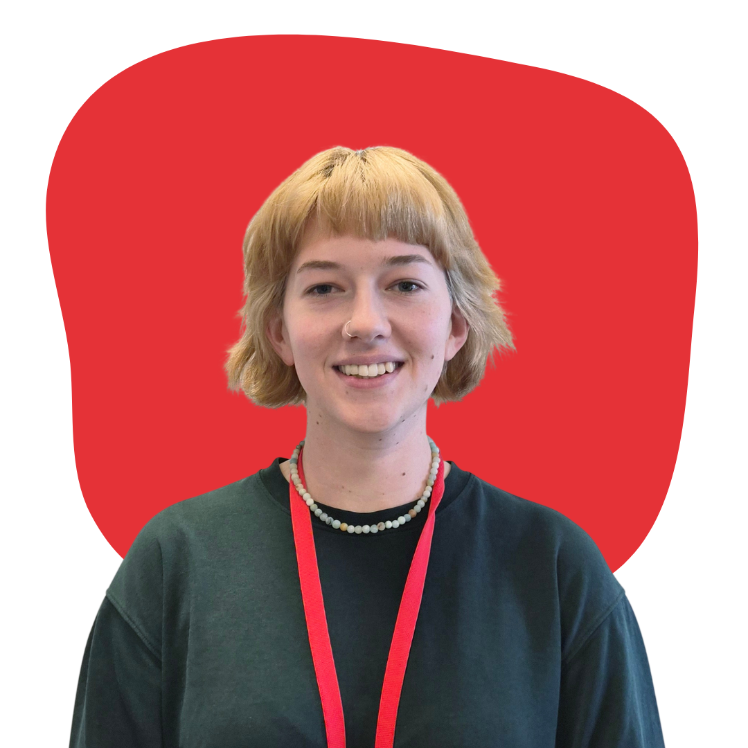 Young woman with short blonde hair, wearing a black shirt, a pearl necklace, and a red lanyard, smiling in front of a red background.