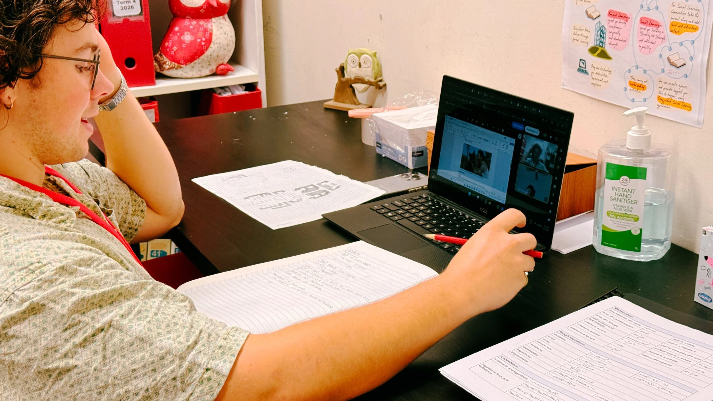 Person sitting at a desk with papers, a laptop, and a hand sanitizer bottle, working on a computer in a classroom or office.