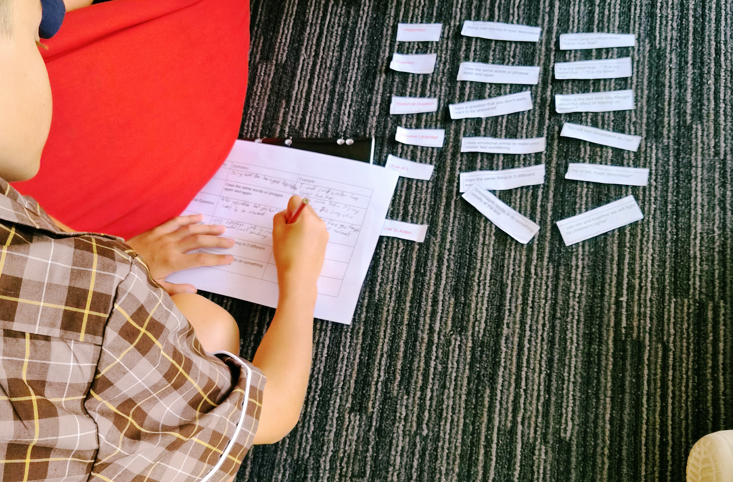 A person sitting on a striped carpet, writing in a notebook. Around them are scattered strips of paper with text, and a larger sheet of paper with handwritten notes.