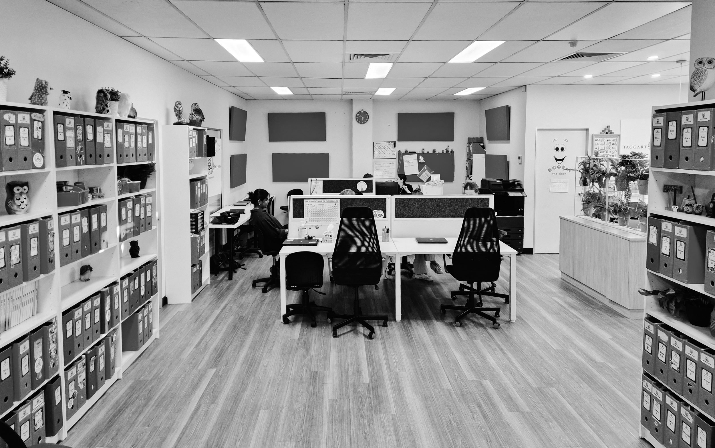 An office with four desks arranged in a square, each with an office chair. There are bookcases filled with binders on both sides, and three people working at their desks. The back wall features three large monitors and a clock. The floor is made of wood, and the ceiling has fluorescent lights.