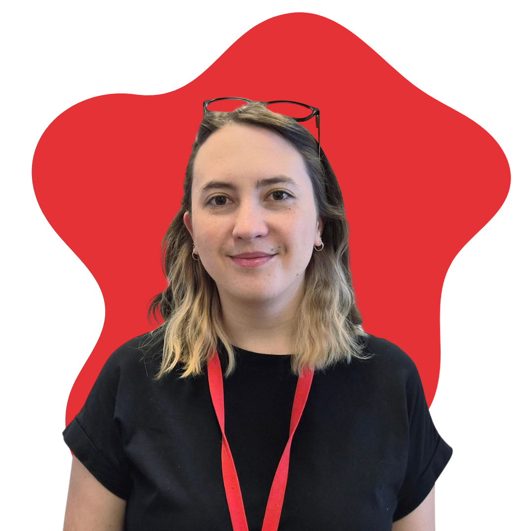 A woman with shoulder-length wavy blonde hair, wearing glasses on her head, hoop earrings, a black T-shirt, and a red lanyard, standing in front of a red background.