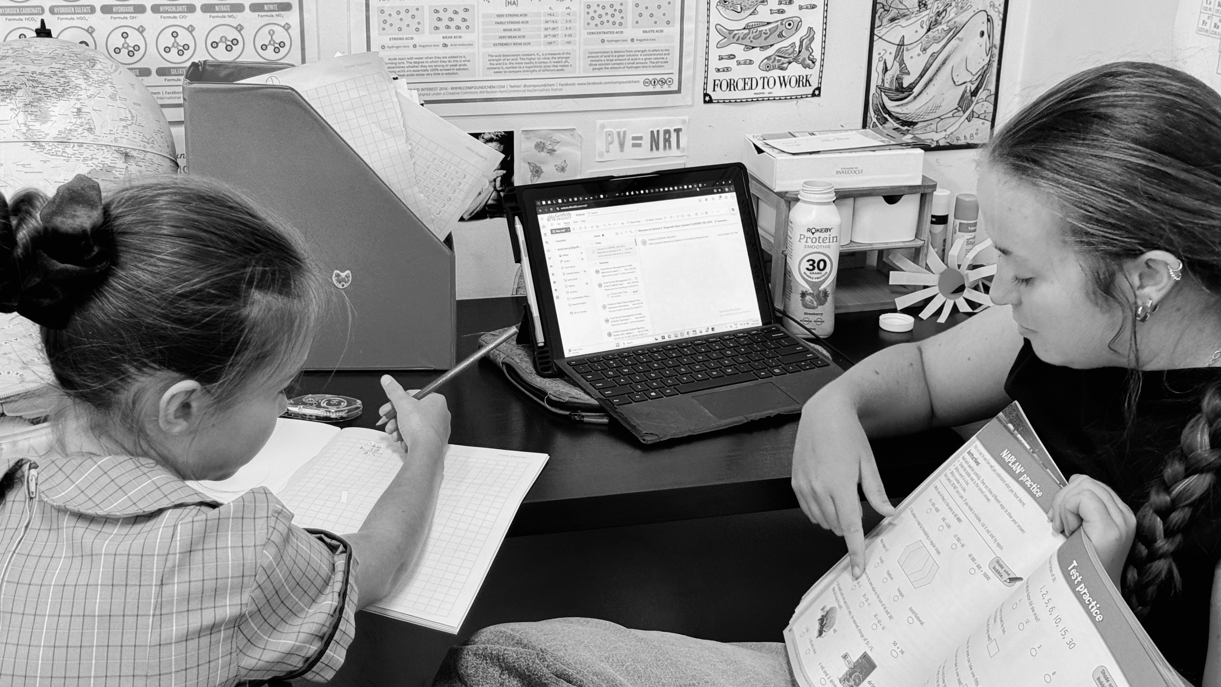 A teacher and a student working at a desk with a laptop, notebooks, and educational materials, in a classroom with posters and classroom supplies on the wall.