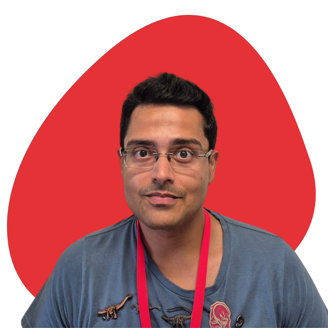 A man with glasses and short dark hair wearing a blue t-shirt with dinosaur patches and a red lanyard, sitting in front of a white background with a large red shape behind him.