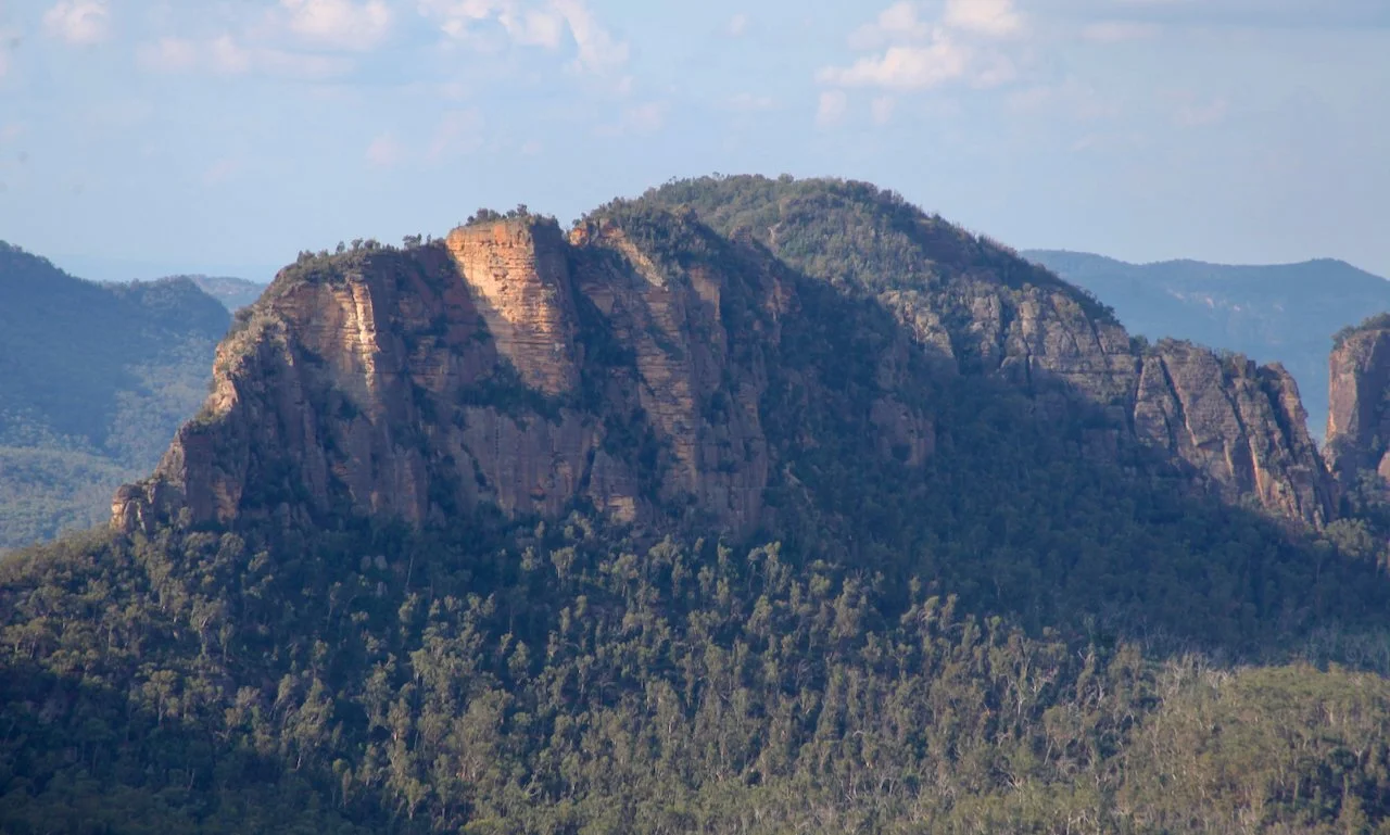 A mountain with rocky cliffs surrounded by dense green forest under a partly cloudy sky.