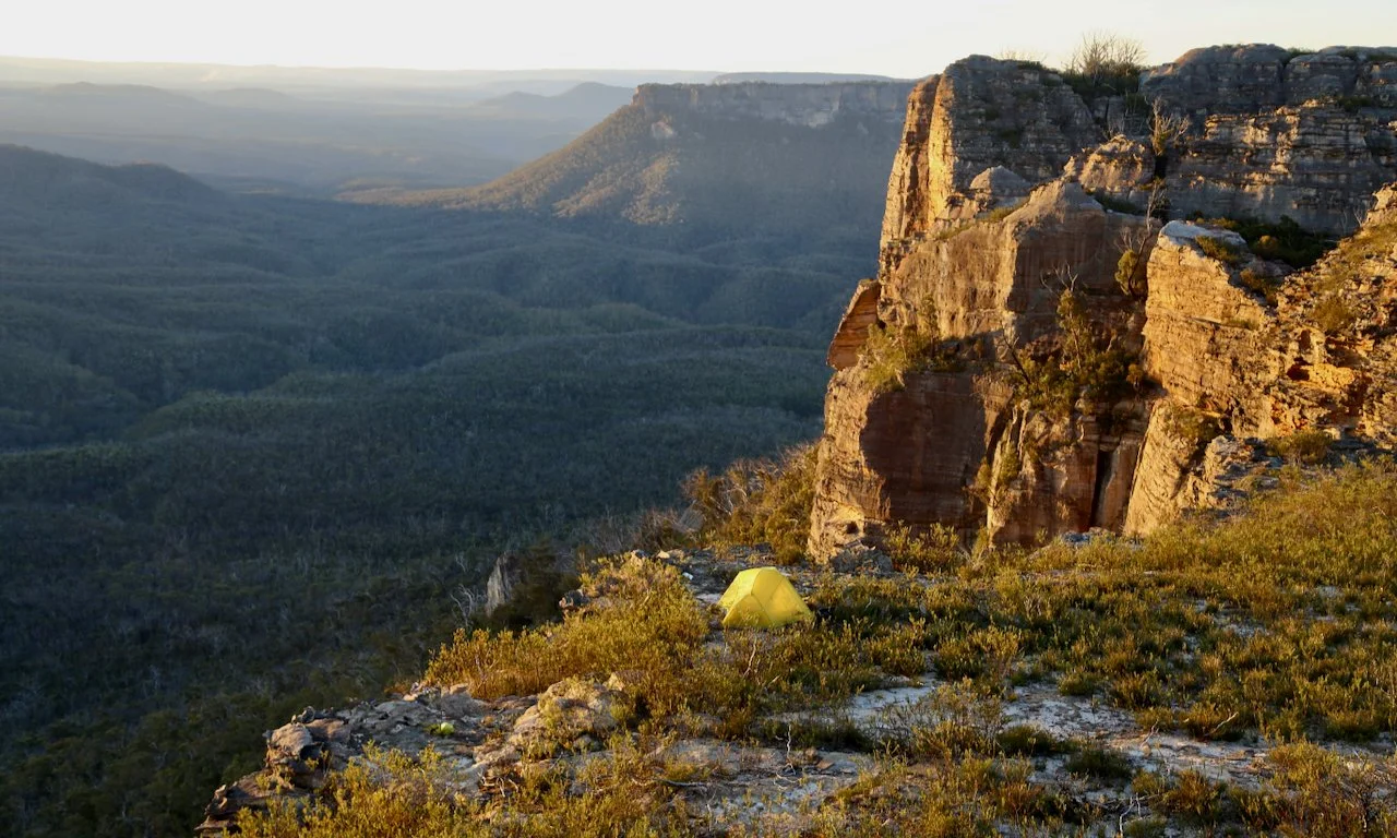 A yellow tent on rocky terrain with green shrubs, surrounded by cliffs and a vast mountain landscape at sunrise or sunset.