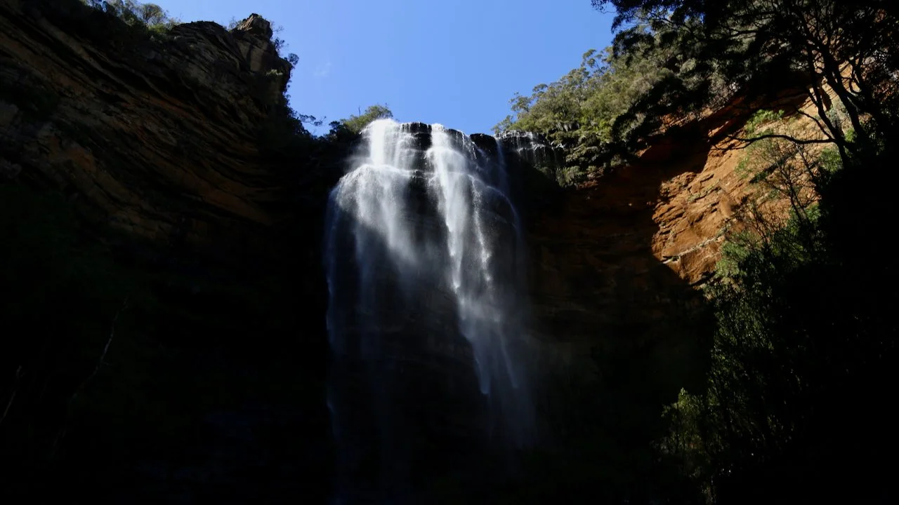 A tall waterfall cascading down a cliff surrounded by trees and rocks, with a blue sky overhead.