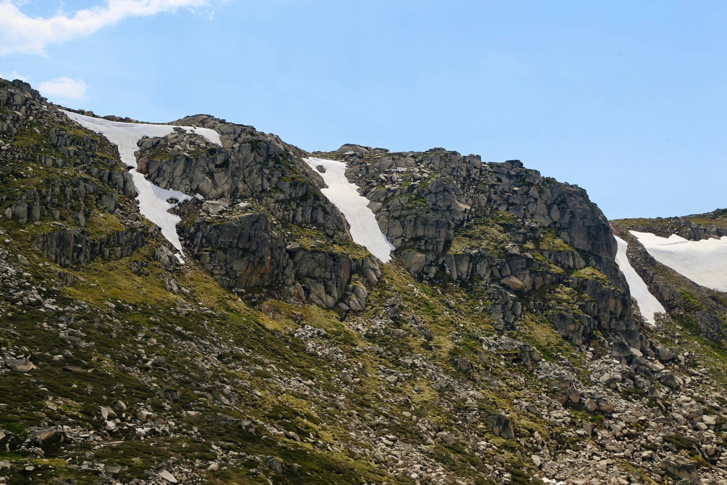Two hikers walking on a snow-covered mountain slope with large rocks and a clear blue sky.