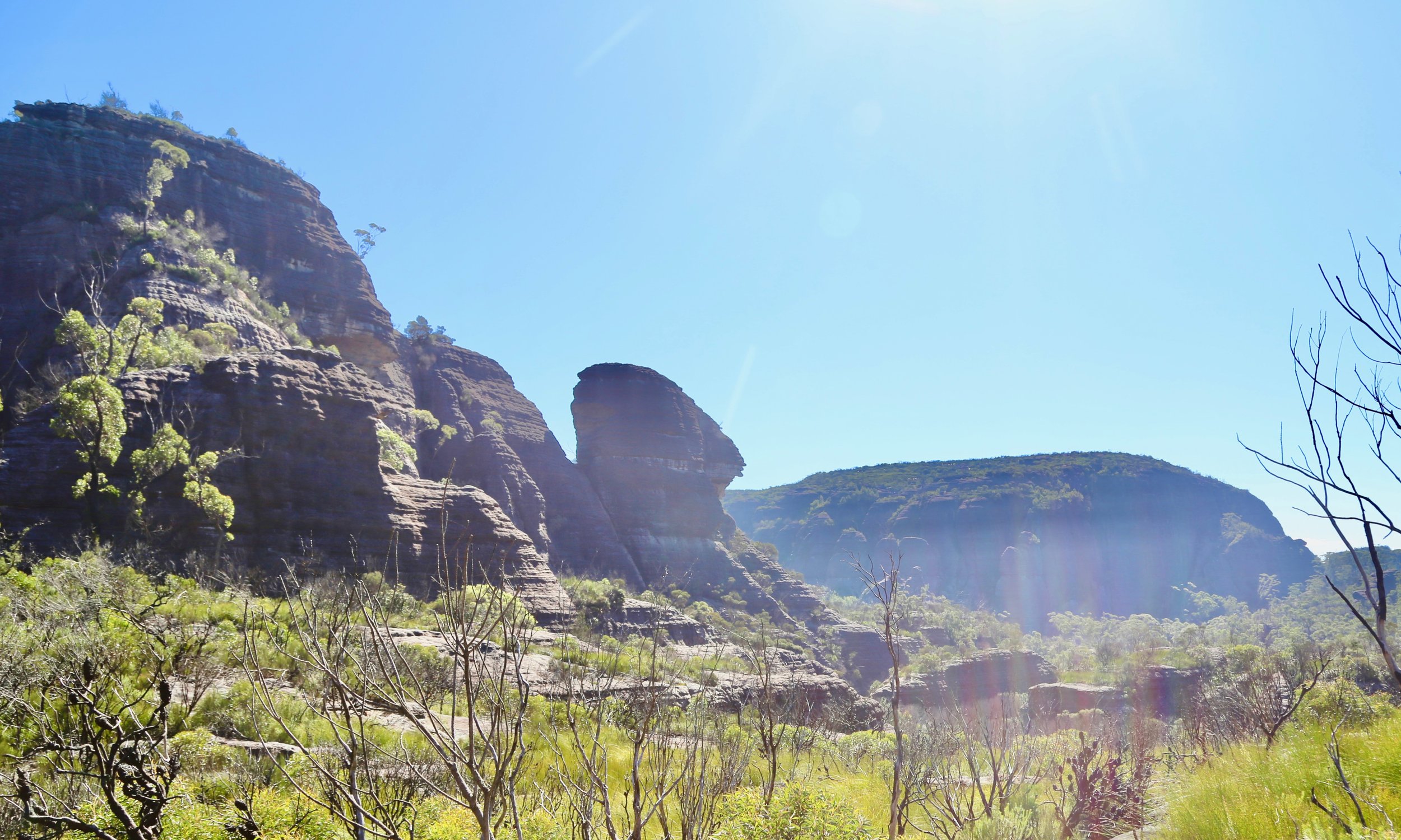 Sunlit rocky mountain landscape with sparse trees and dry vegetation in the foreground, clear blue sky with lens flare.