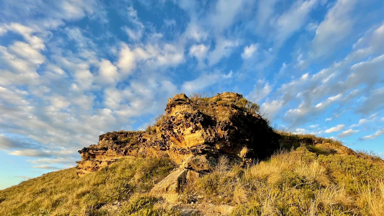 A rocky hill with sparse vegetation under a cloudy blue sky.