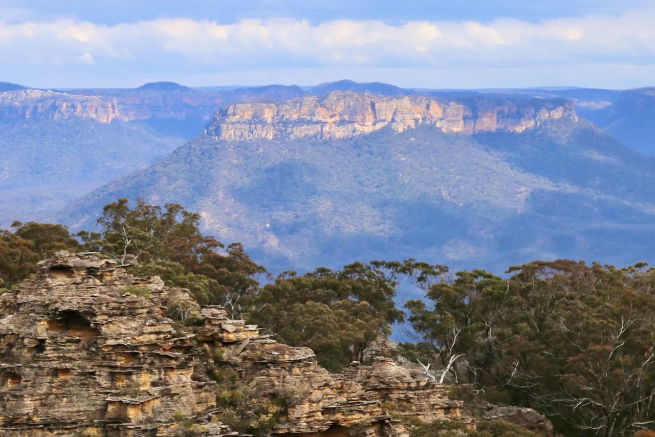 Scenic view of layered rock formations in the foreground, dense trees in the midground, and a blue mountain range under a cloudy sky in the background.