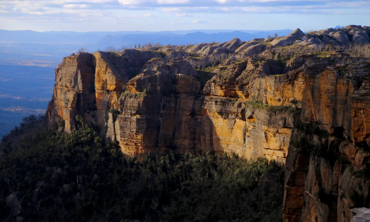 A scenic view of a rugged mountain cliff with layered rock formations and sparse vegetation, with distant mountains in the background.