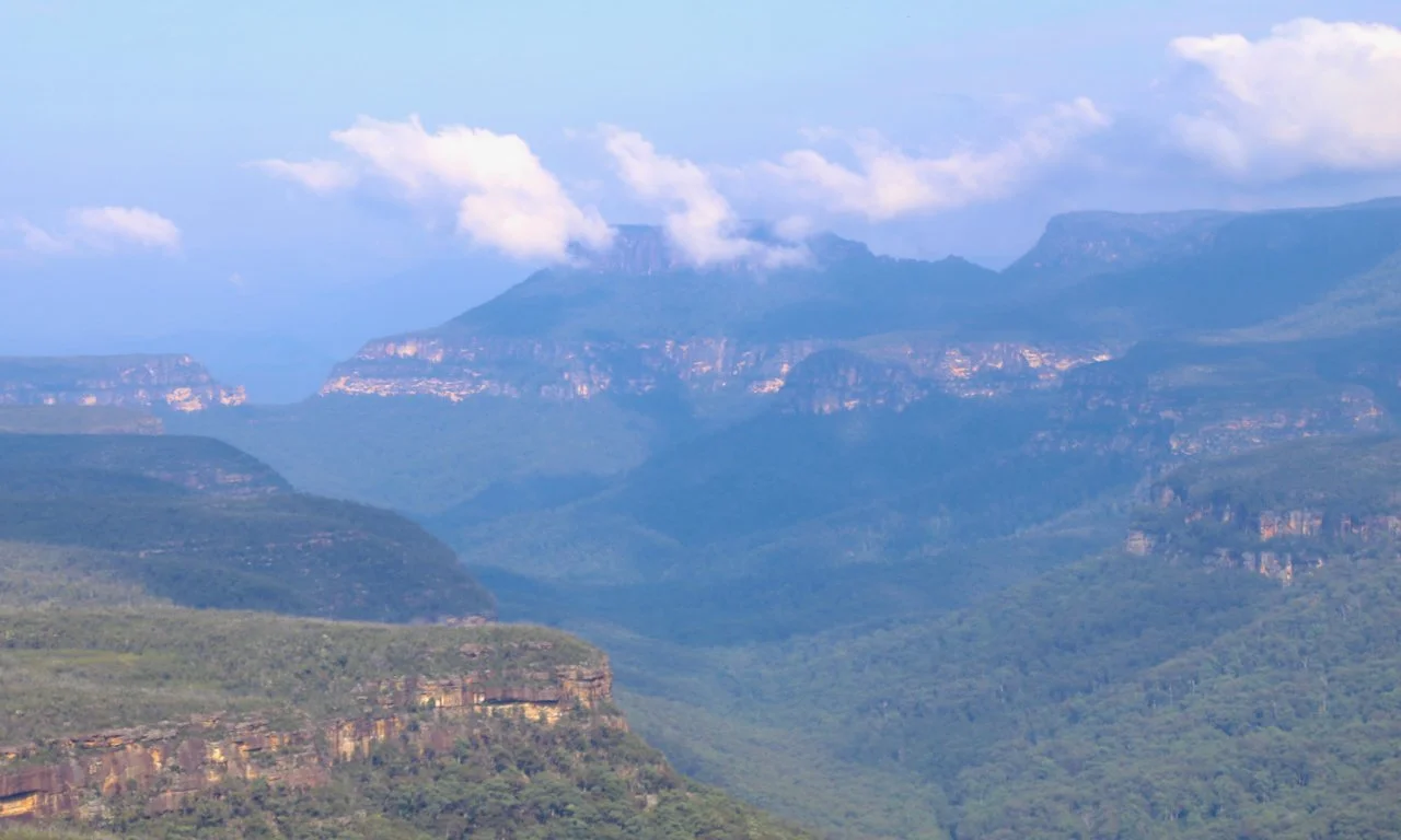 Scenic view of blue mountain range with clouds above, green forests on the slopes, and rugged cliffs.