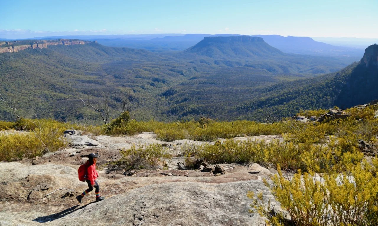 Hiker with a red backpack and black hat walking on rocky terrain in a mountain landscape with green valleys and plateaued mountains in the background.