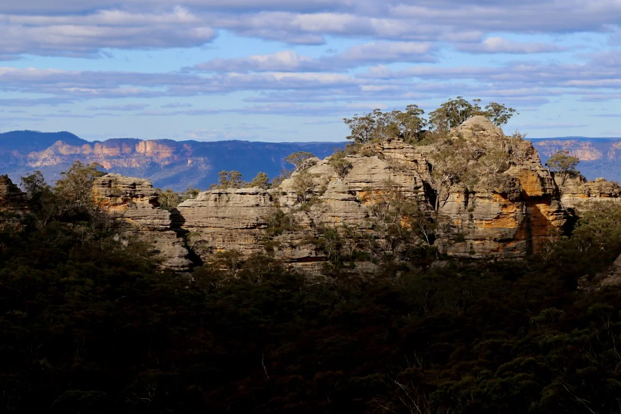 Rock formations and trees in a natural landscape with mountains in the background and a partly cloudy sky.