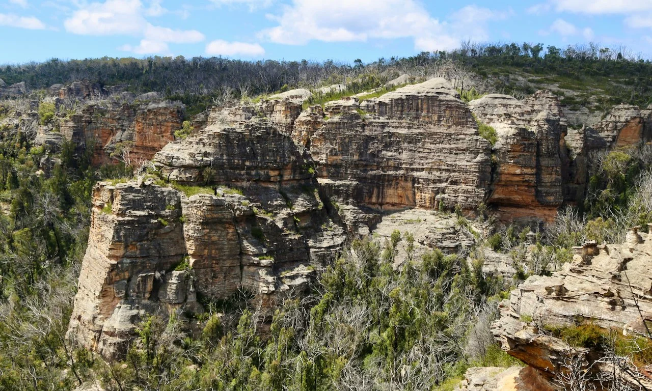 Rock formations and cliffs in a canyon with sparse vegetation and a partly cloudy sky.
