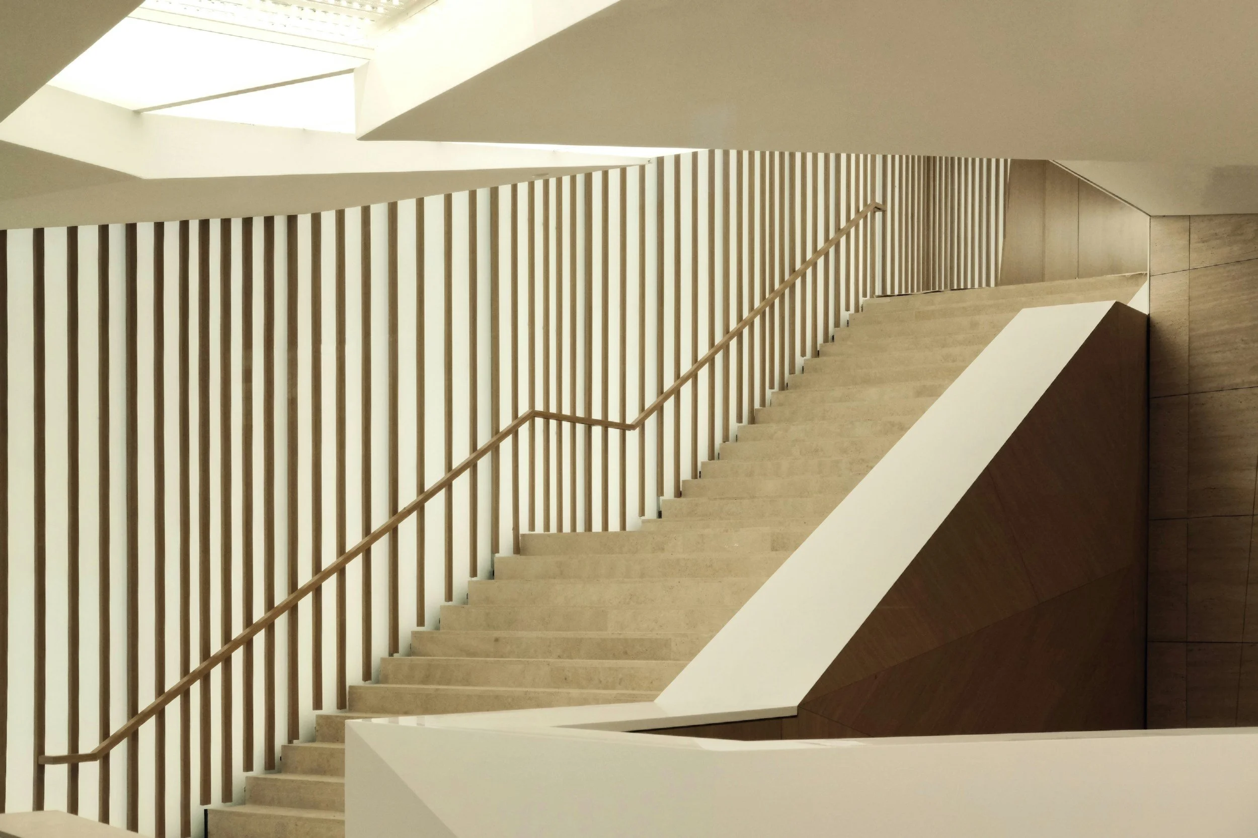 An indoor staircase viewed from above, with beige steps, a wooden handrail, and vertical wooden slats as a safety barrier. The staircase is enclosed by walls and natural light is coming from a window or skylight at the top.