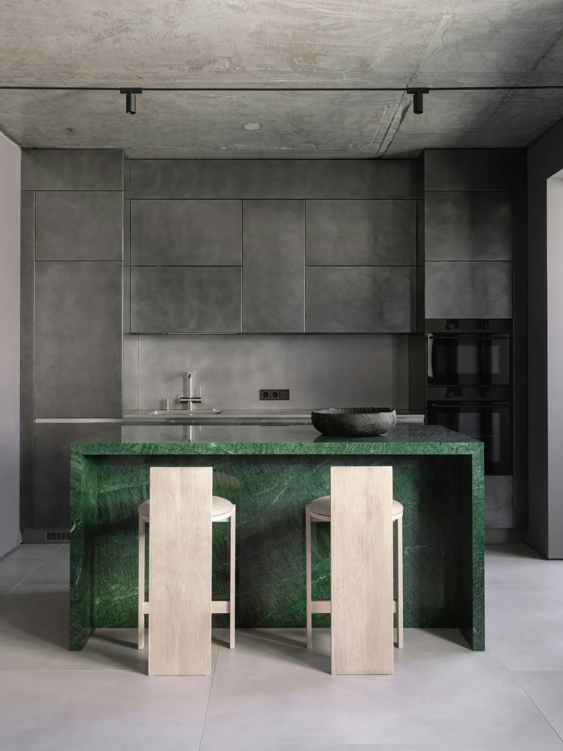 Modern kitchen with dark gray cabinets, a black oven, a green marble kitchen island with two beige chairs, and a concrete ceiling.