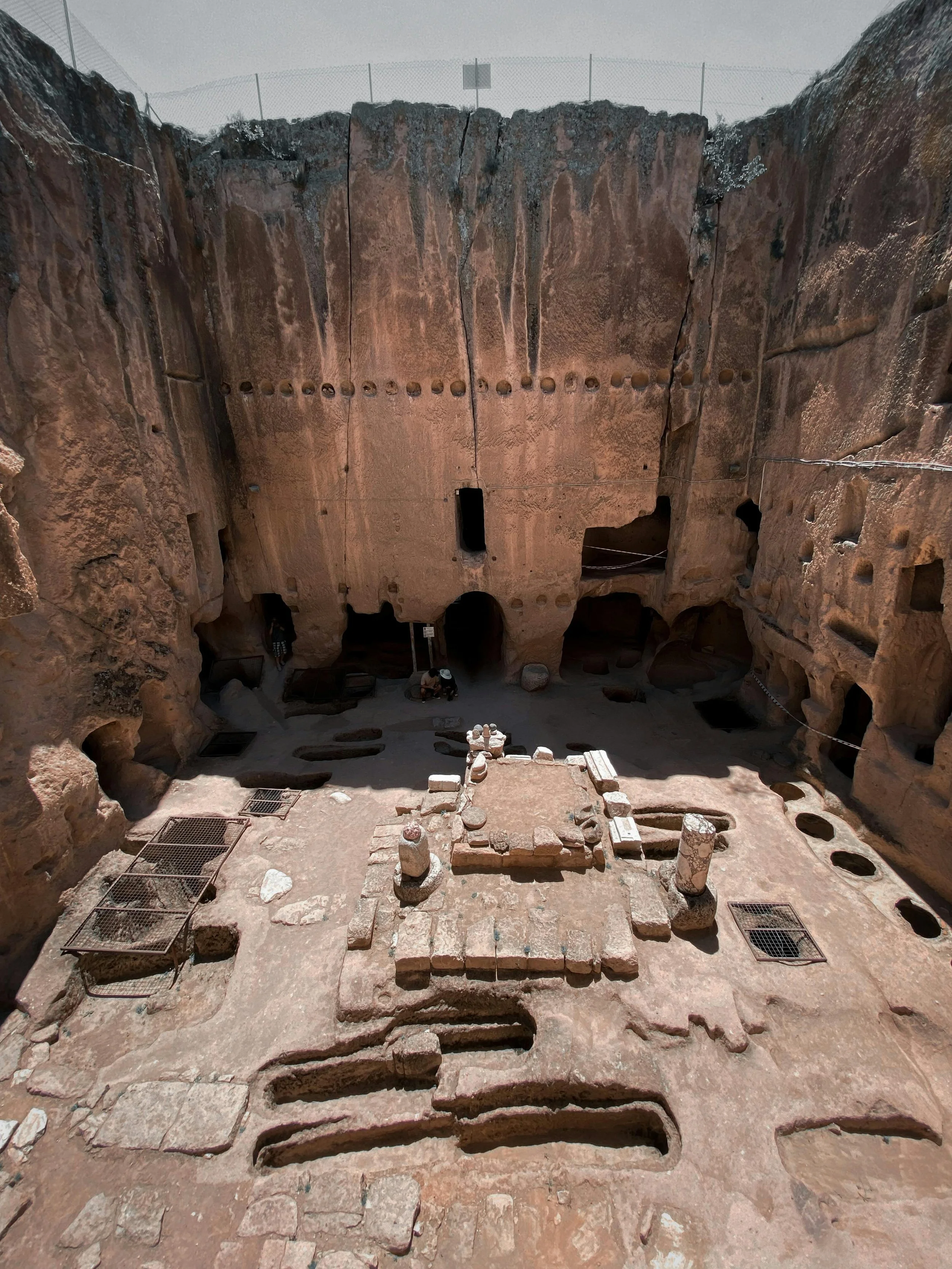 An ancient archaeological site with a sunken central area surrounded by tall stone walls and steps leading down into the site, with a partially intact stone altar or monument at the center.