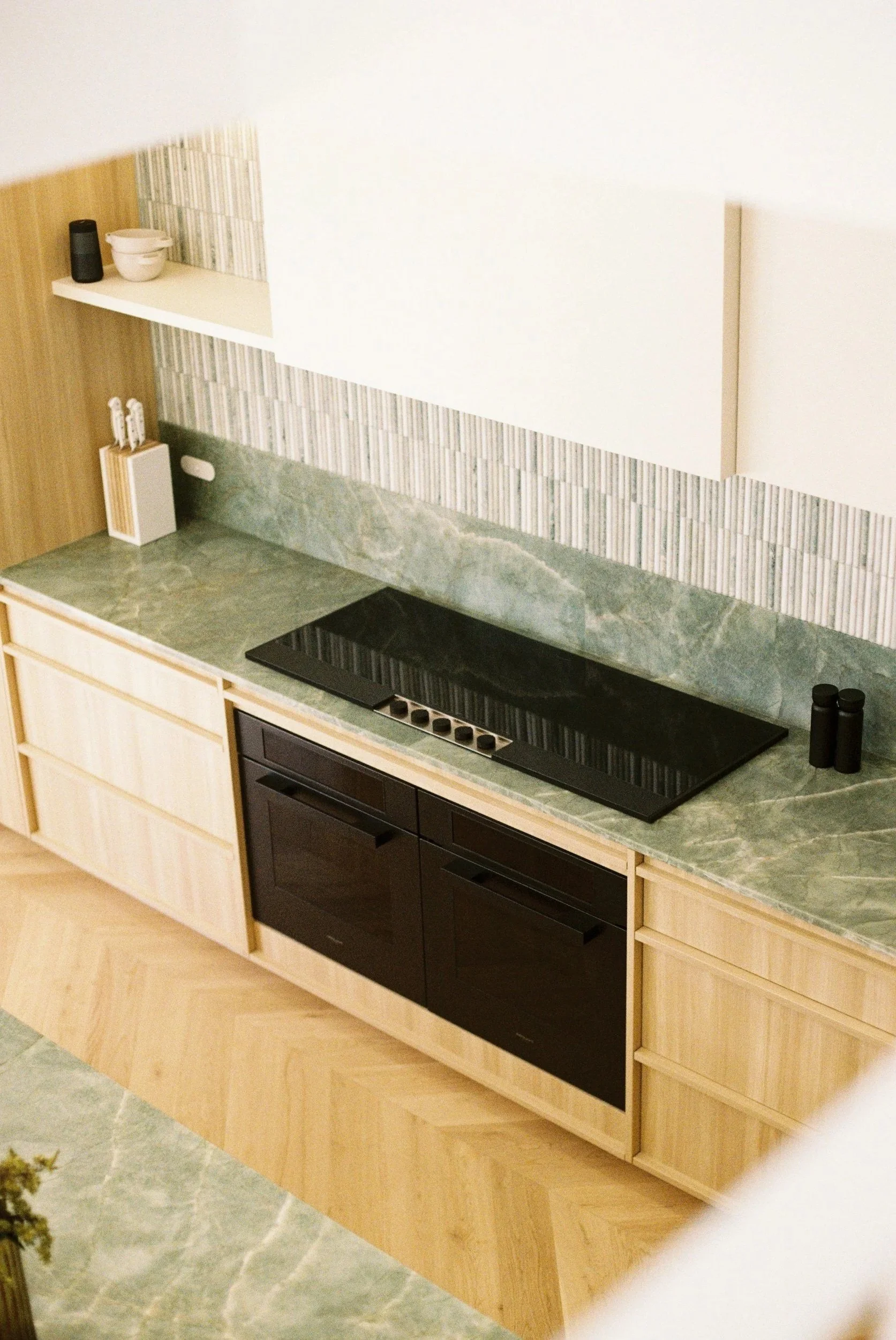 Modern kitchen with green marble countertops, light wood cabinets, black stove, and minimal decorative items.
