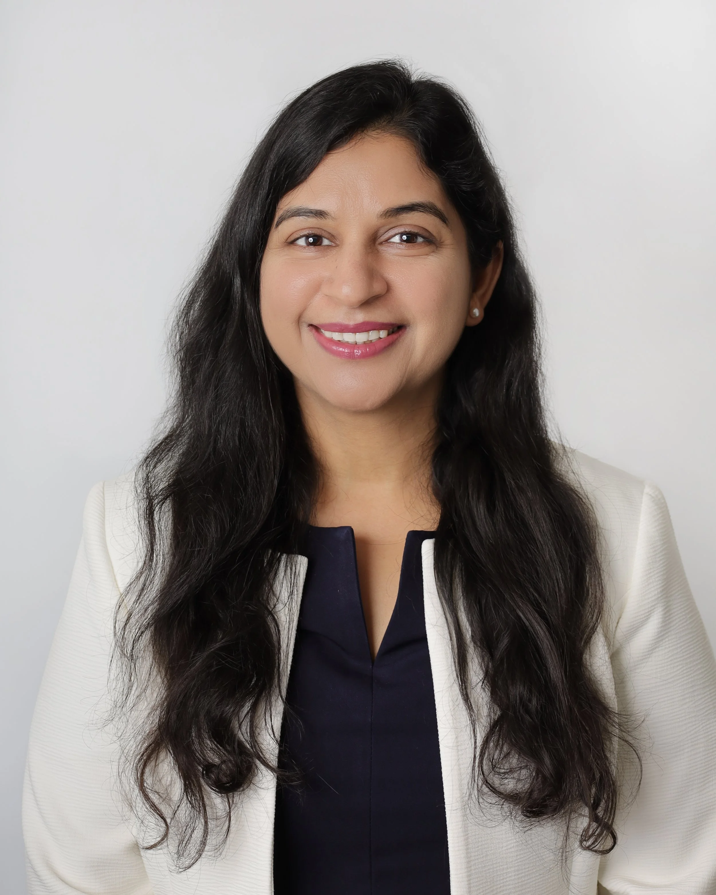 Portrait of a smiling woman with long dark hair, wearing a cream blazer and navy top, against a plain white background.