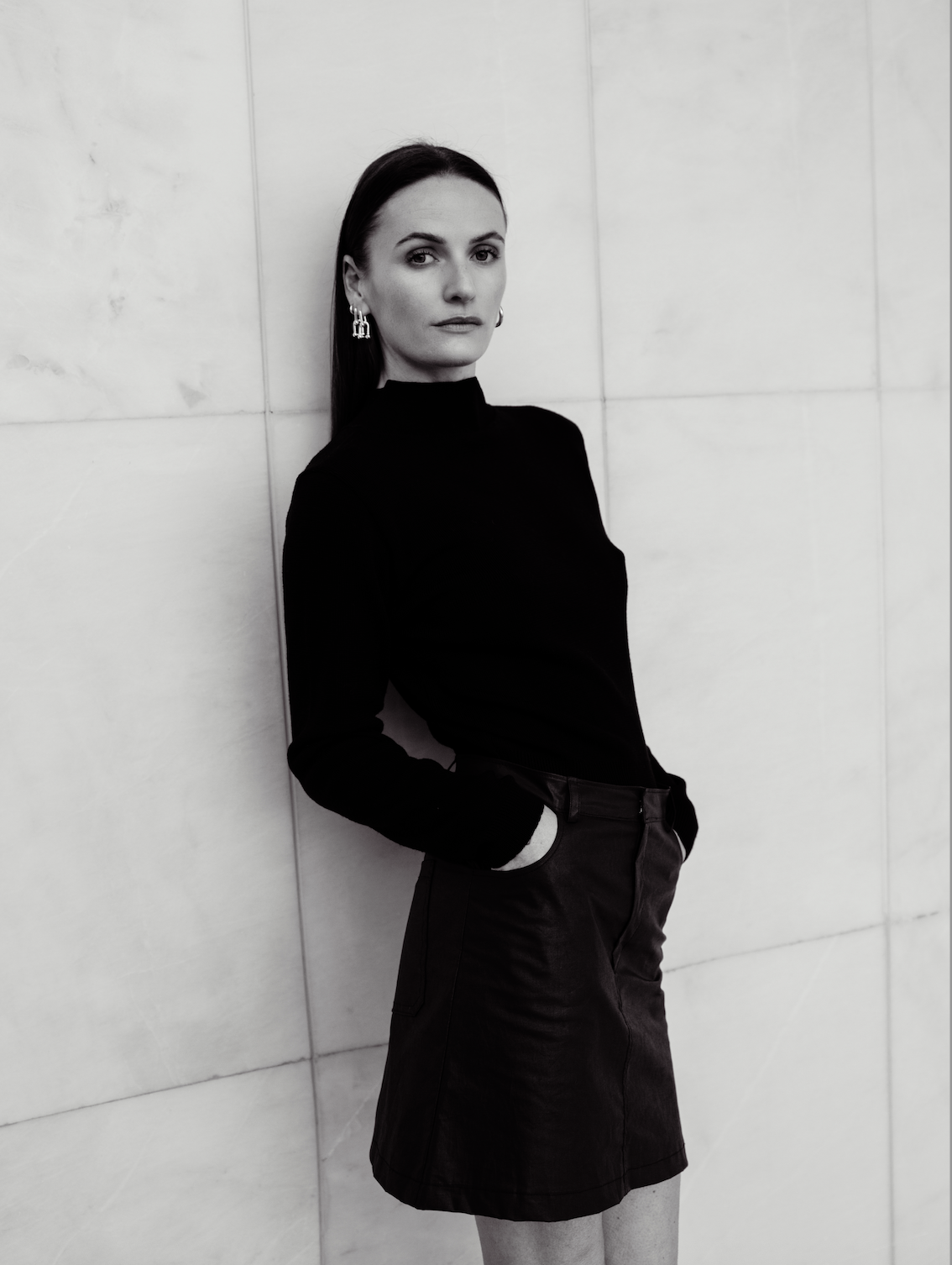 Une femme en skateboard noir et blanc, portant un haut noir à col haut, une jupe noire, et des boucles d'oreilles pendantes, se tenant contre un mur en carrelage.