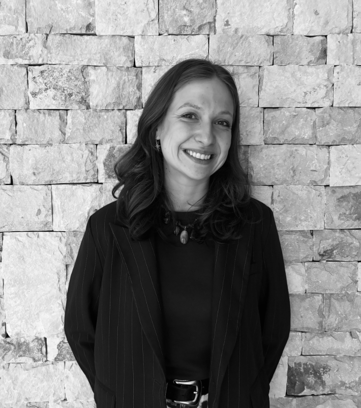 Une femme souriante avec des cheveux ondulés foncés, portant un blazer à rayures et un collier avec une pierre, se tenant devant un mur en pierre.