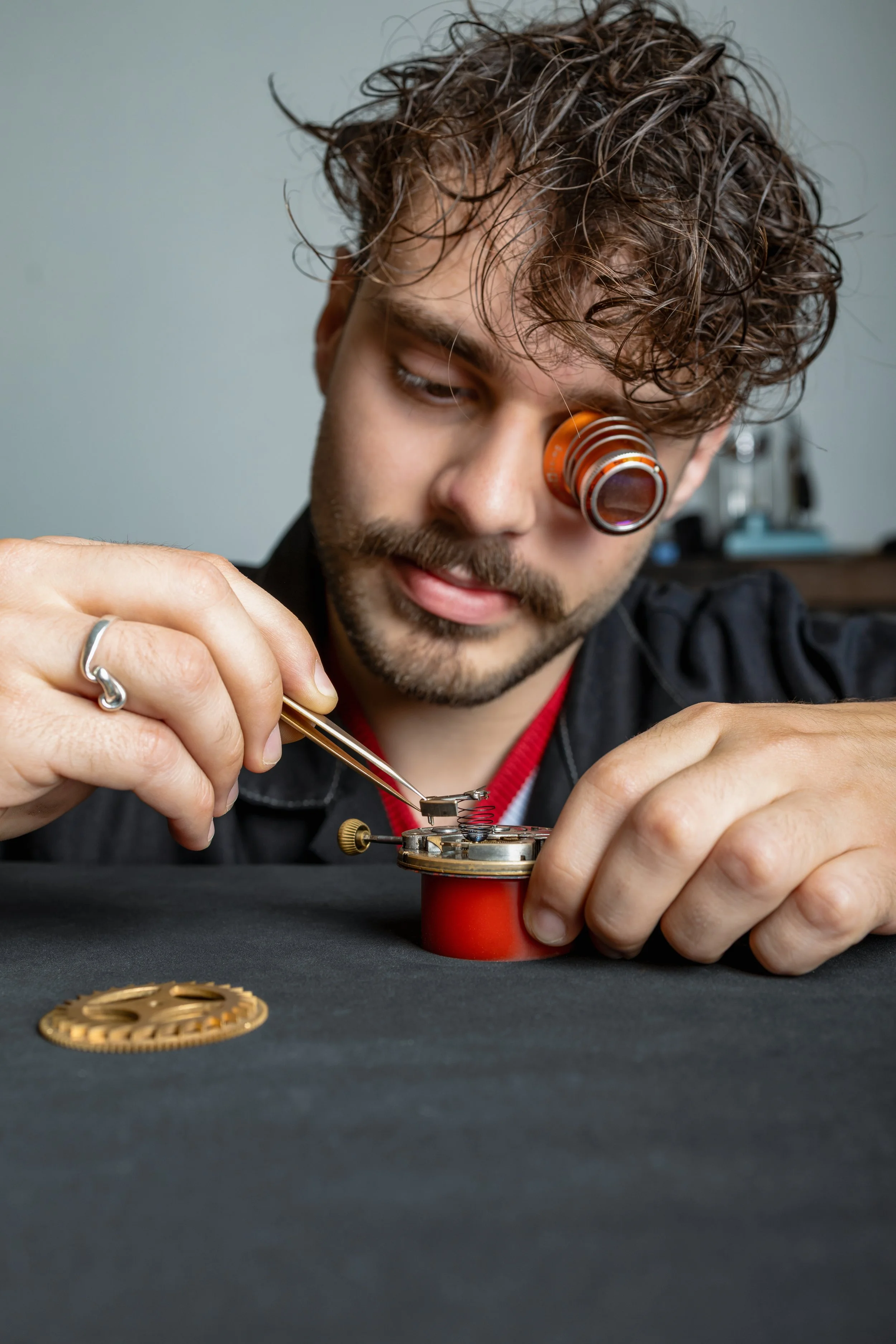 Un homme effectue une réparation horlogère, utilisant une loupe sur son œil et des outils pour manipuler un mécanisme de montre.
