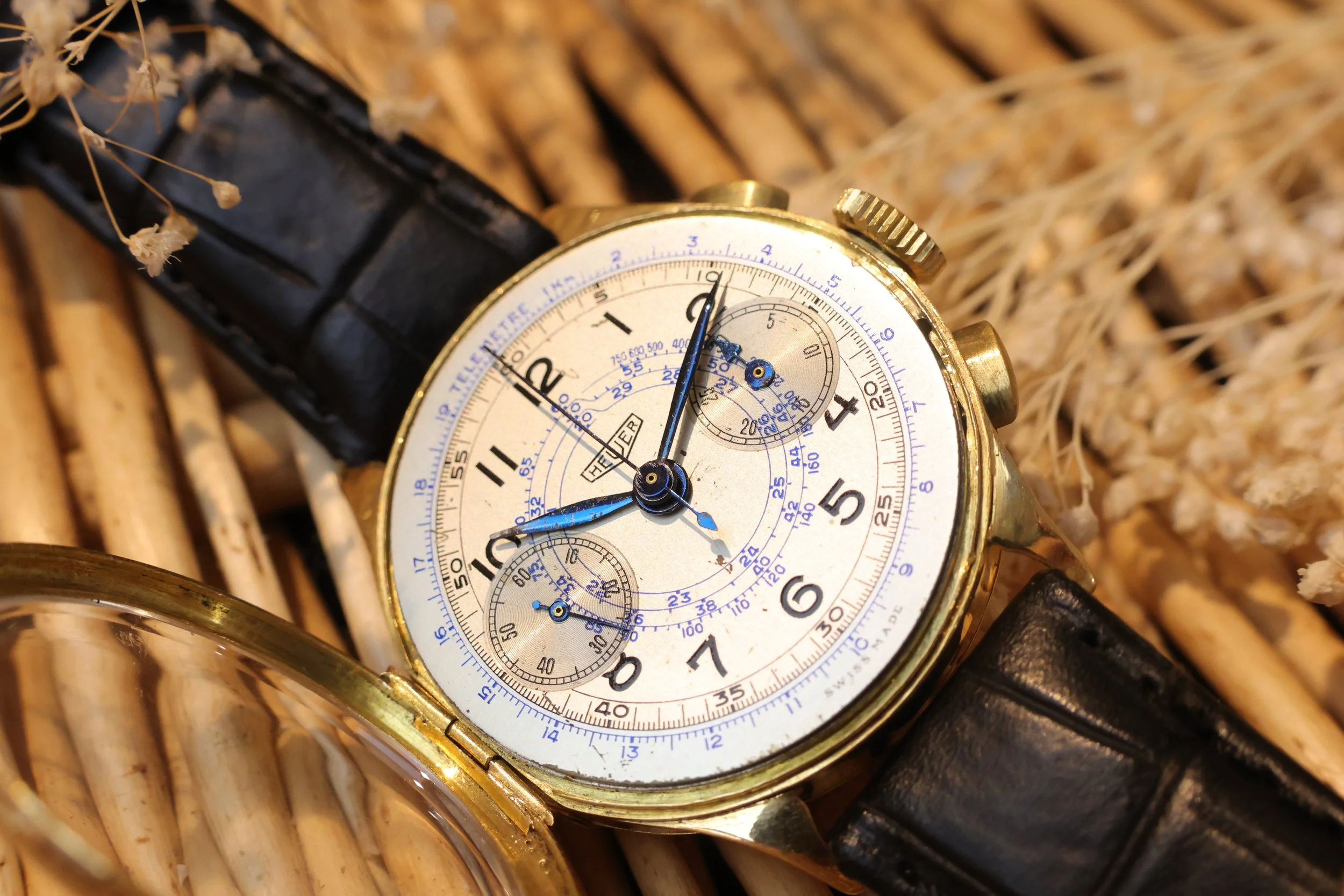 Montre ancienne avec un fond blanc, chiffres arabes noirs, et plusieurs sous-cadrans, enroulée dans un bracelet en cuir noir, posée sur une surface en bois ou osier.