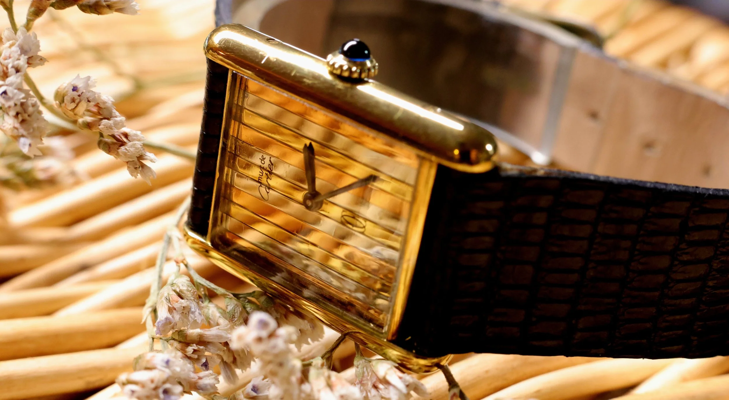 Montre vintage avec un bracelet en cuir noir, placée sur un panier en osier avec des fleurs séchées à côté.