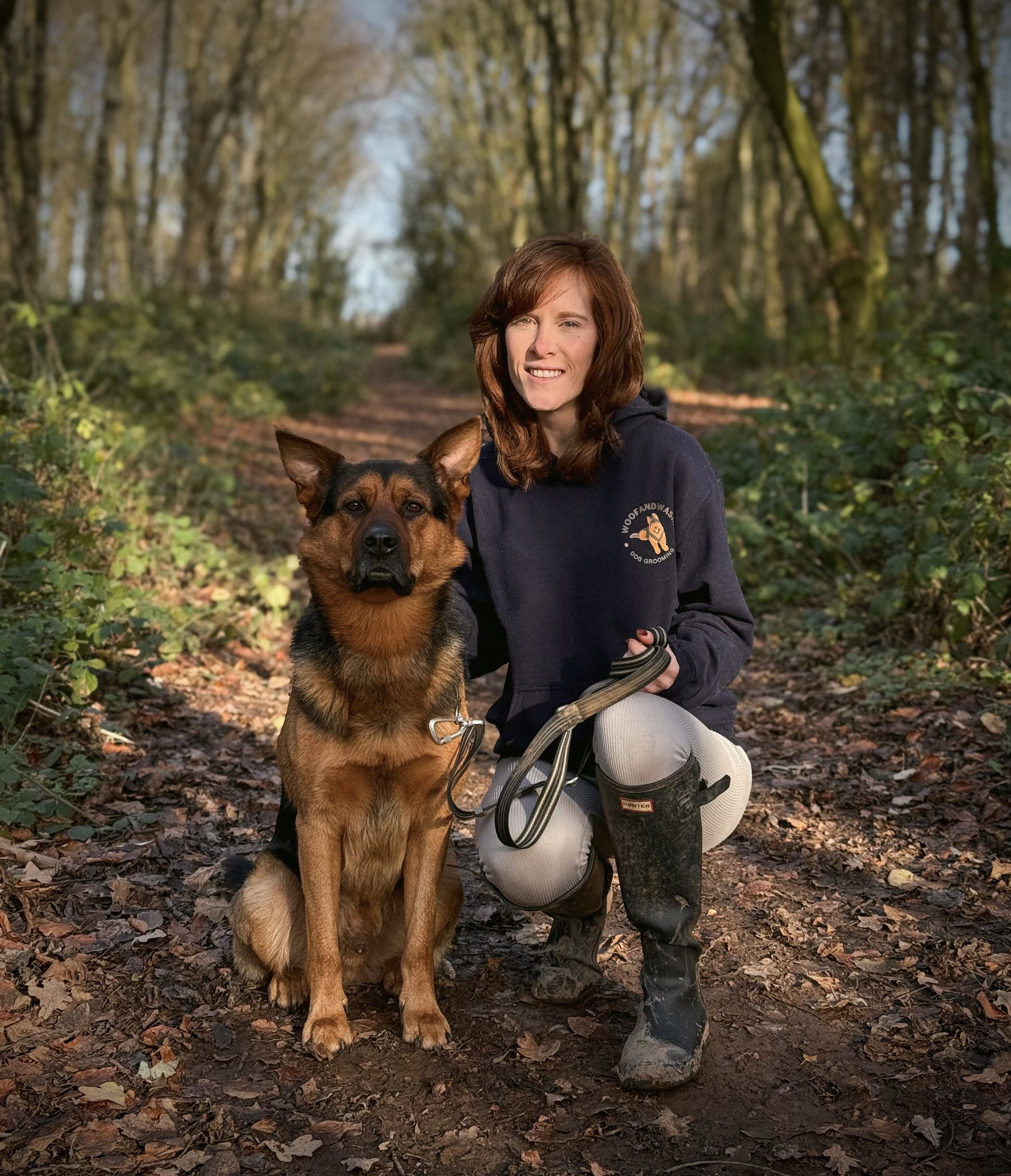 A woman crouching next to a dog in a wooded trail, both looking at the camera, during the daytime.
