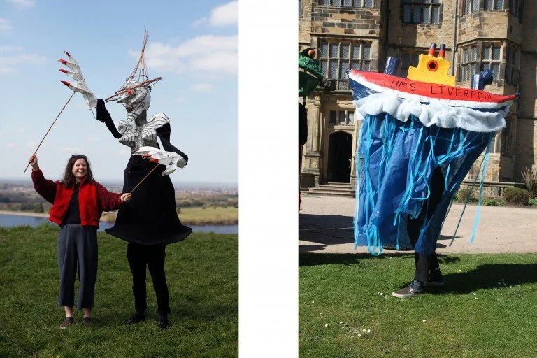 Two people hold kites designed as a dragon and a ship, standing on a grassy field with a river in the background.