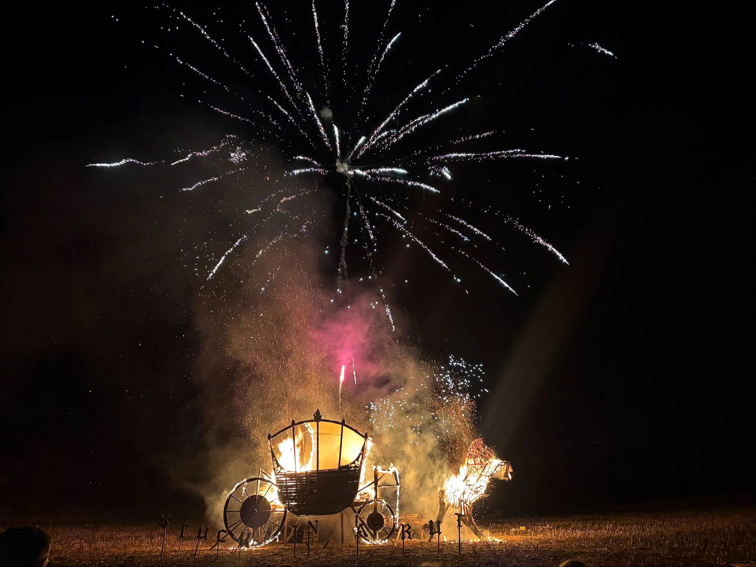 Fireworks explode in the night sky above a large, wooden, wagon-shaped structure that is burning, with flames visible at the top. The scene suggests a celebration or festival.