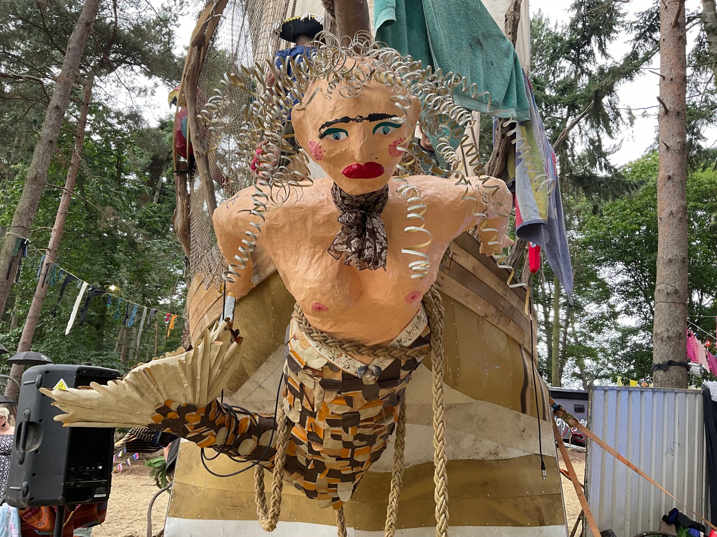 A large colorful puppet of a woman with curly blonde hair, blue eye makeup, and bright red lipstick, hanging on a wooden structure in a forested area.