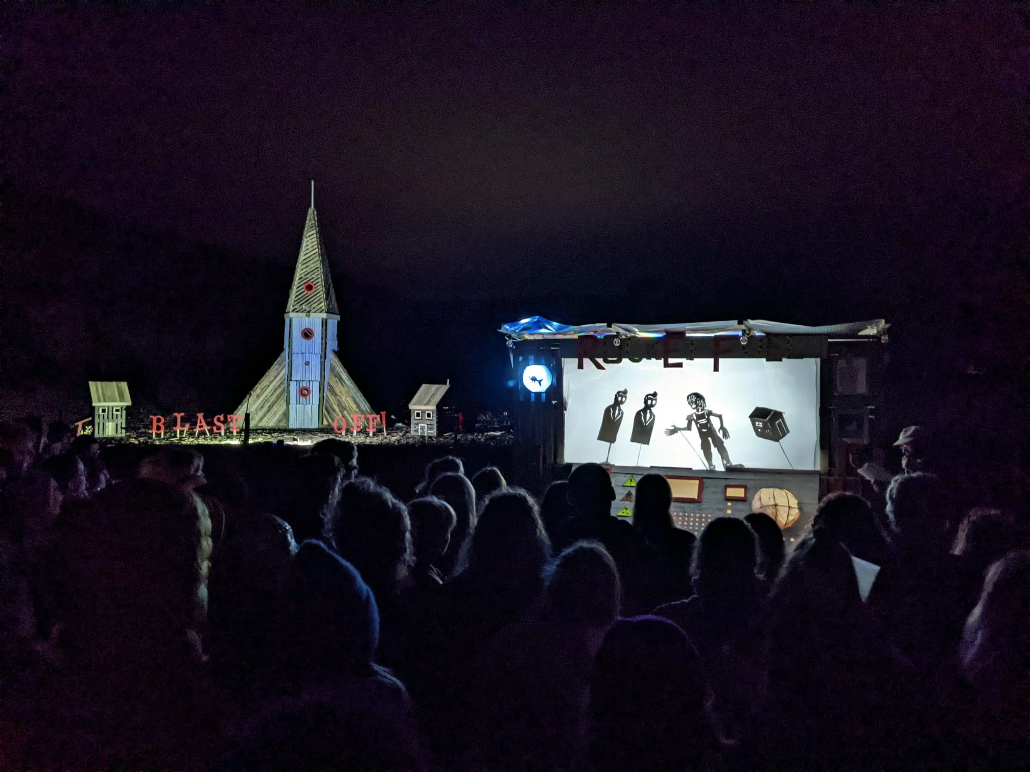 Nighttime scene of an outdoor theater with audience members watching a puppet show on a stage with silhouettes of puppet characters and a backdrop. Decorative structures and a tall church steeple are visible in the background.