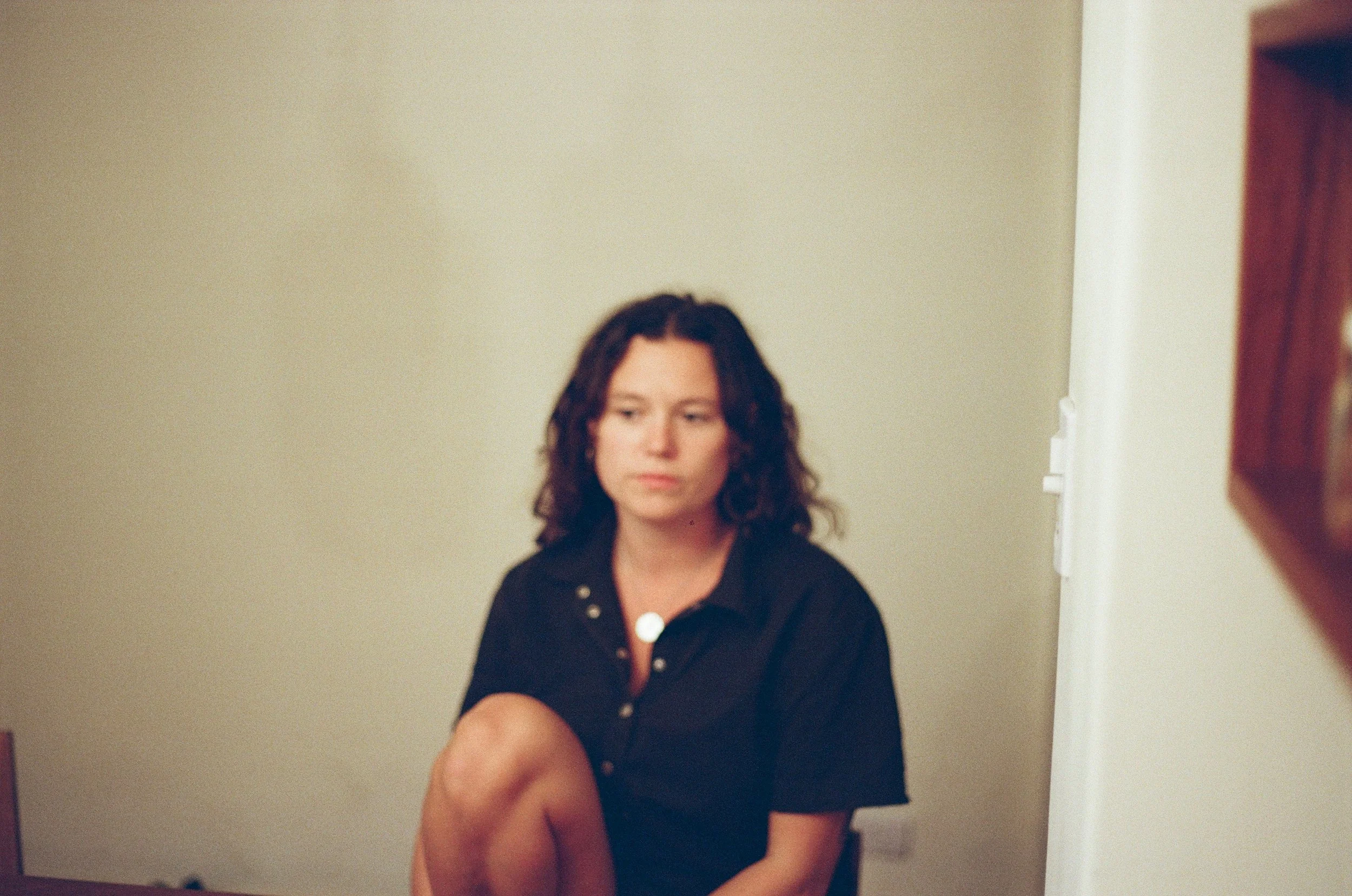 A woman with wavy brown hair wearing a black shirt sitting against a beige wall.
