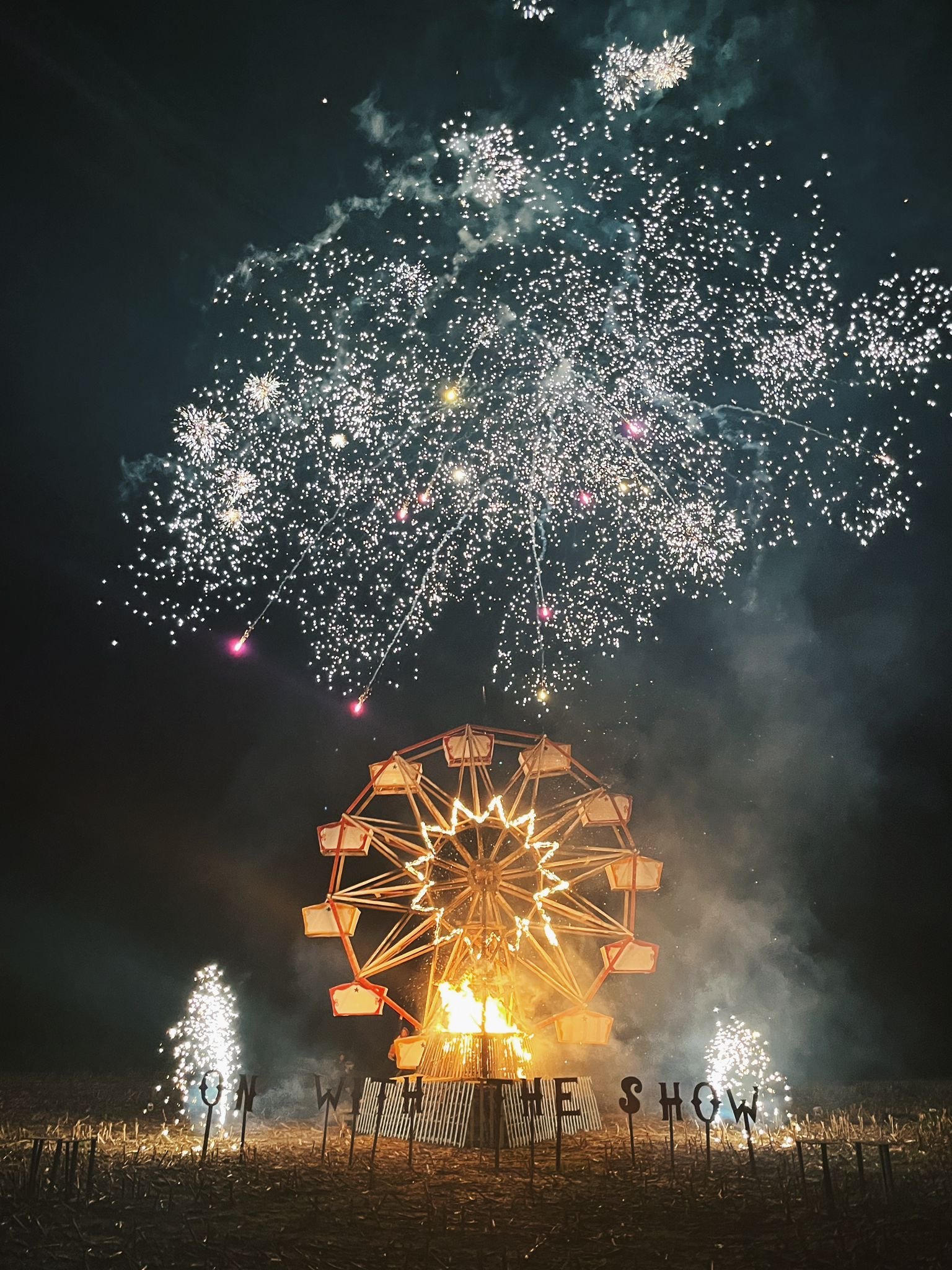 A Ferris wheel lit up at night with fireworks exploding in the sky above.