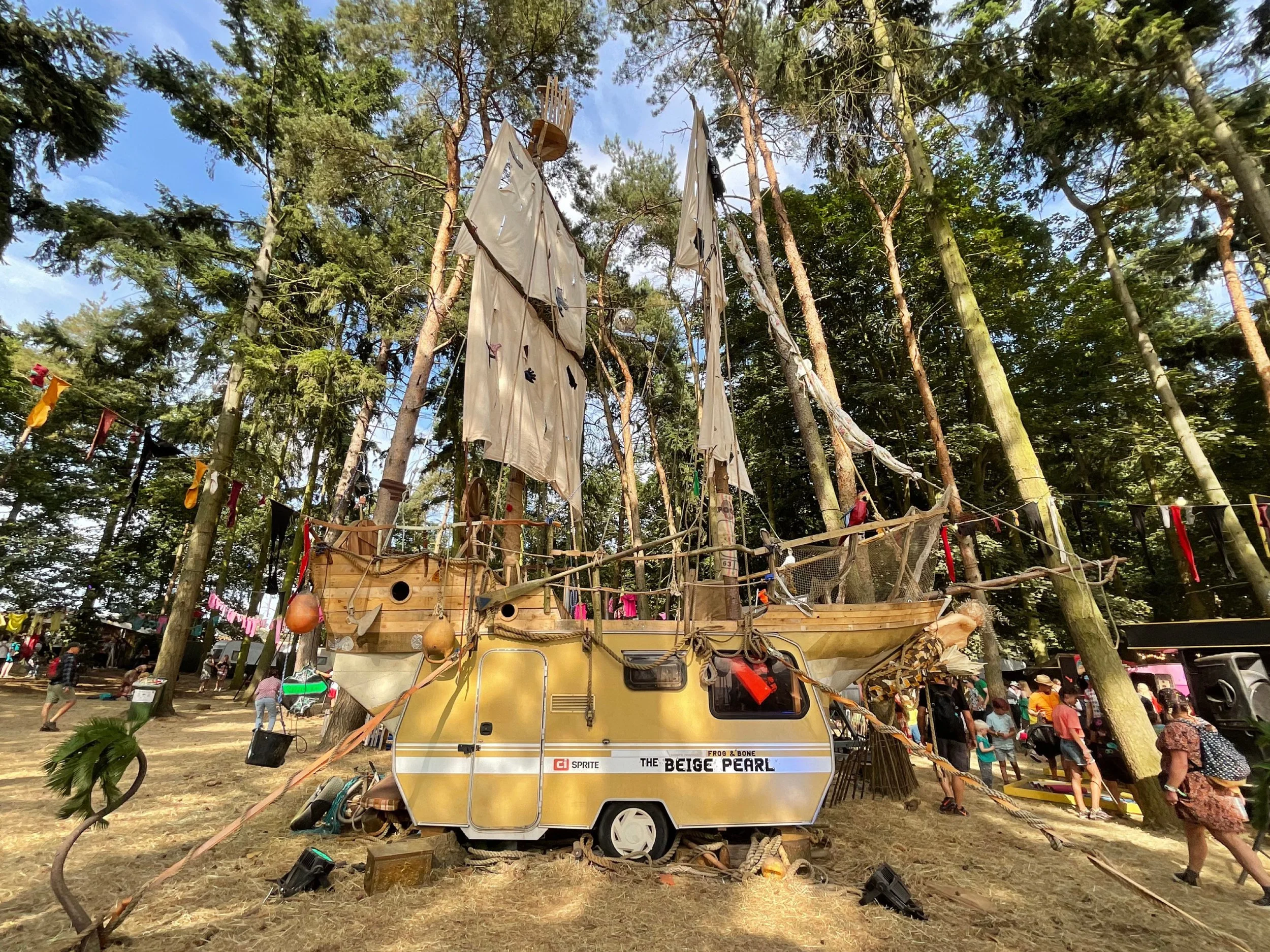 A large yellow and brown pirate ship-themed mobile structure, resembling a boat, is decorated with various nautical accessories and materials, surrounded by people and trees in a park or fair setting.