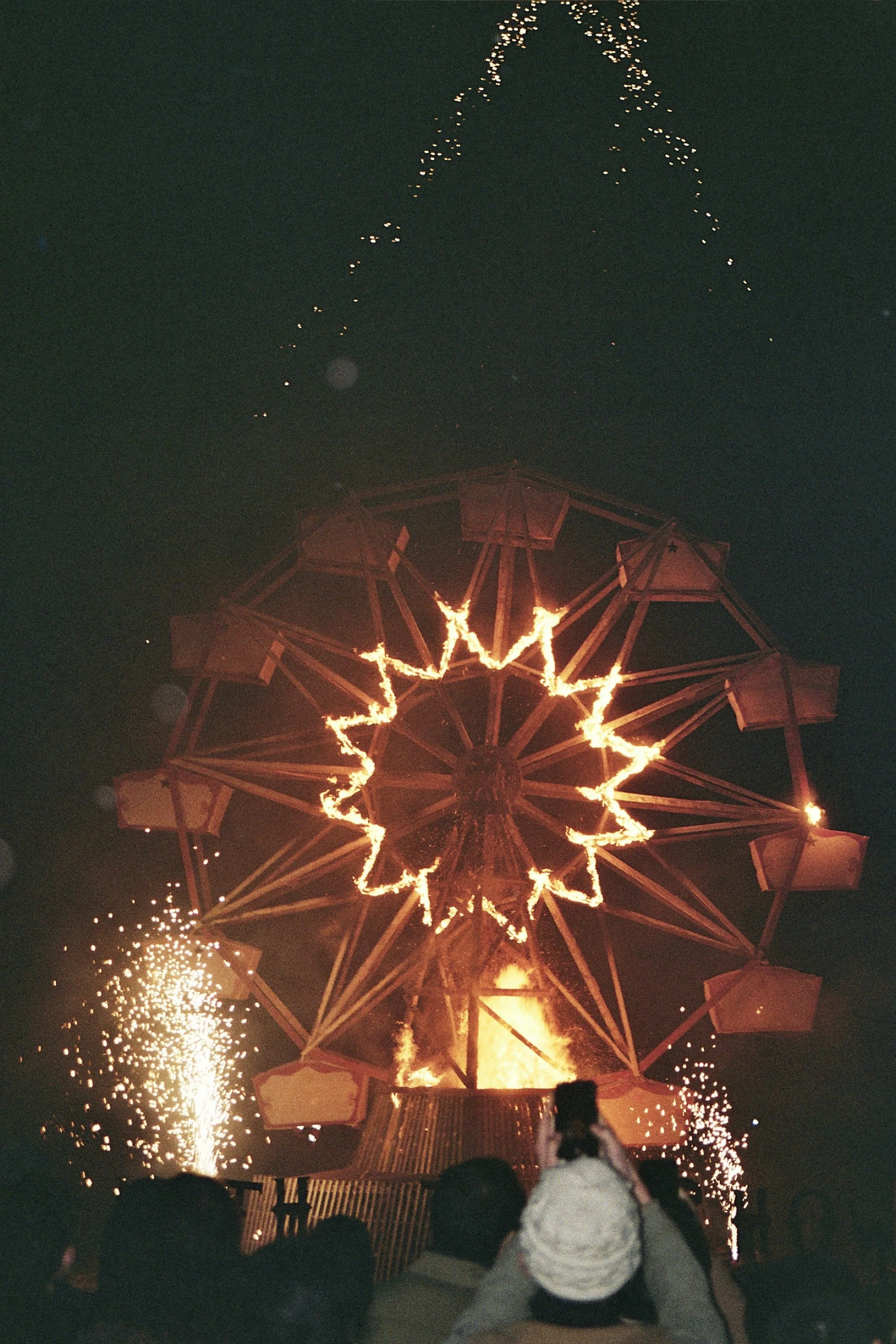 Fireworks display with bright sparks and light patterns lighting up the night sky, people watching the show, with one person taking a photo or video, and large geometric structure illuminated at the bottom