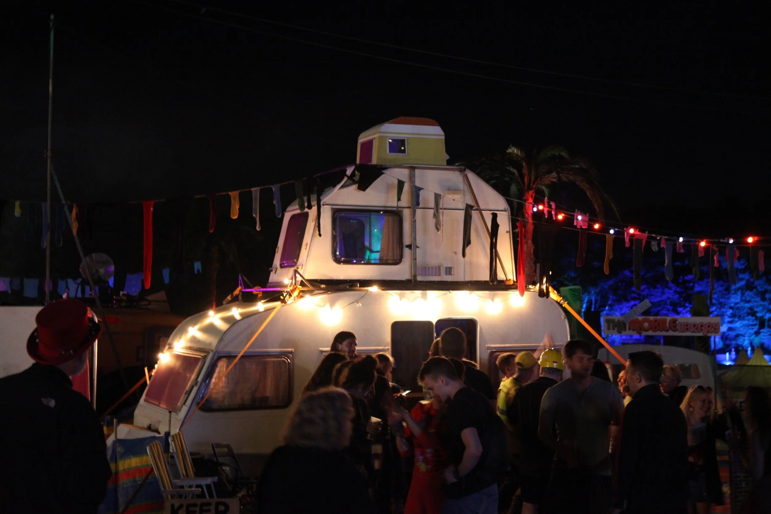 Night scene at a fair or carnival with an old camper trailer decorated with colorful lights and hanging fabric strips, surrounded by people and illuminated by string lights, with some palm trees and a sign in the background.