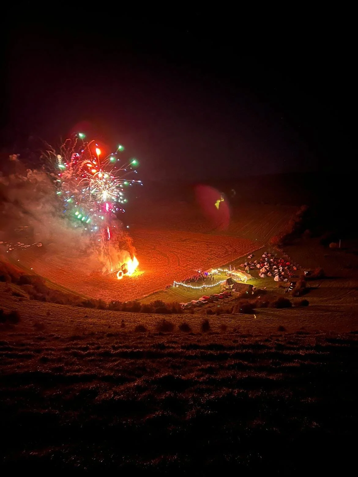 Nighttime scene of fireworks exploding over a rural landscape with tents, cars, and a large bonfire on a grassy hill.