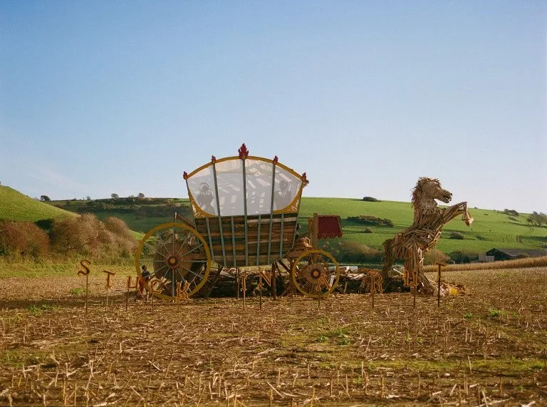 A large sculpture of a fairy tale-style carriage with a horse made of straw or similar material, set in an open field with rolling green hills in the background.