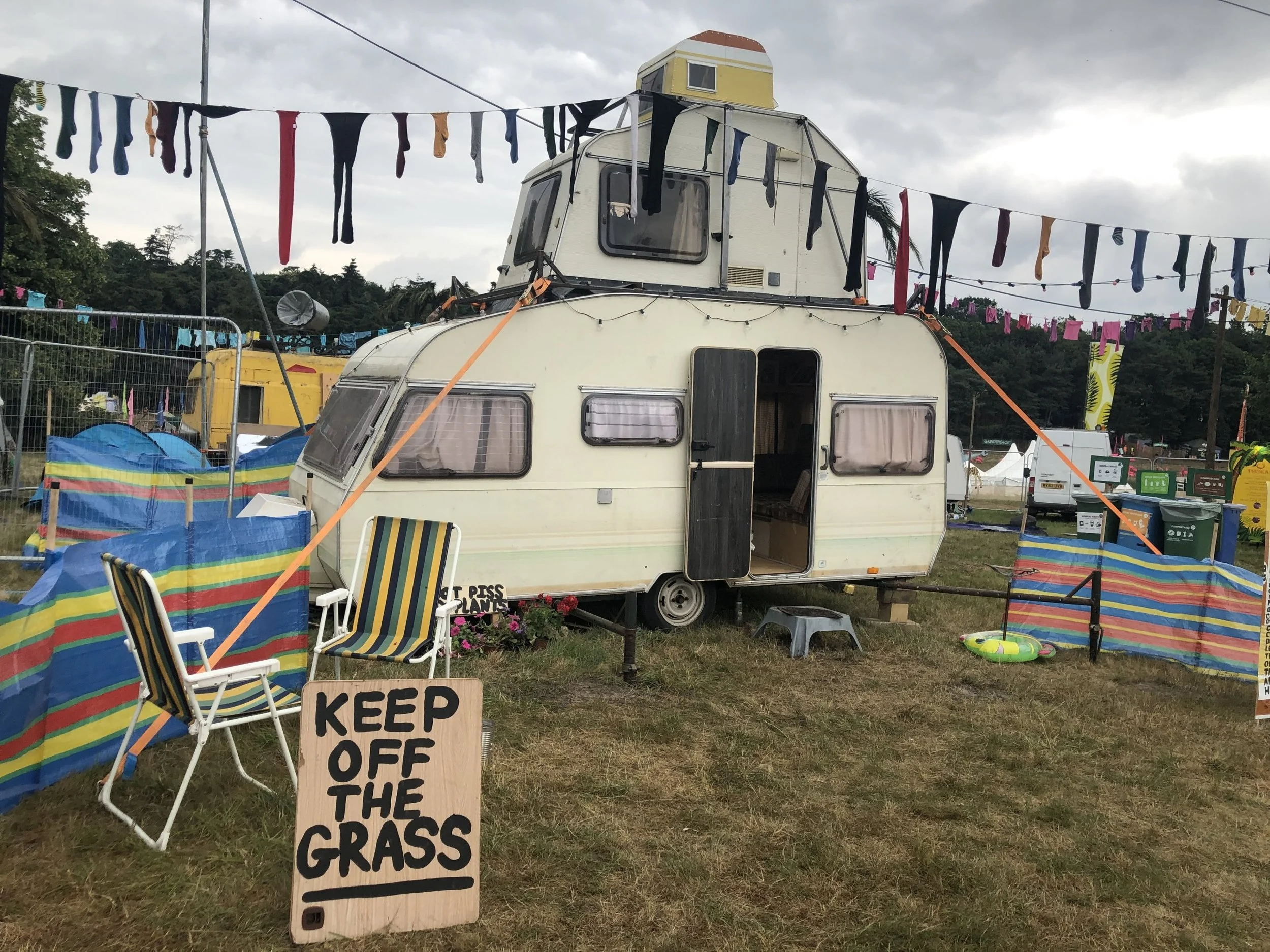 A vintage camper trailer decorated with string lights and surrounded by colorful chairs and a sign that says 'Keep Off the Grass.' In the background, laundry hangs on lines, and the sky is cloudy.