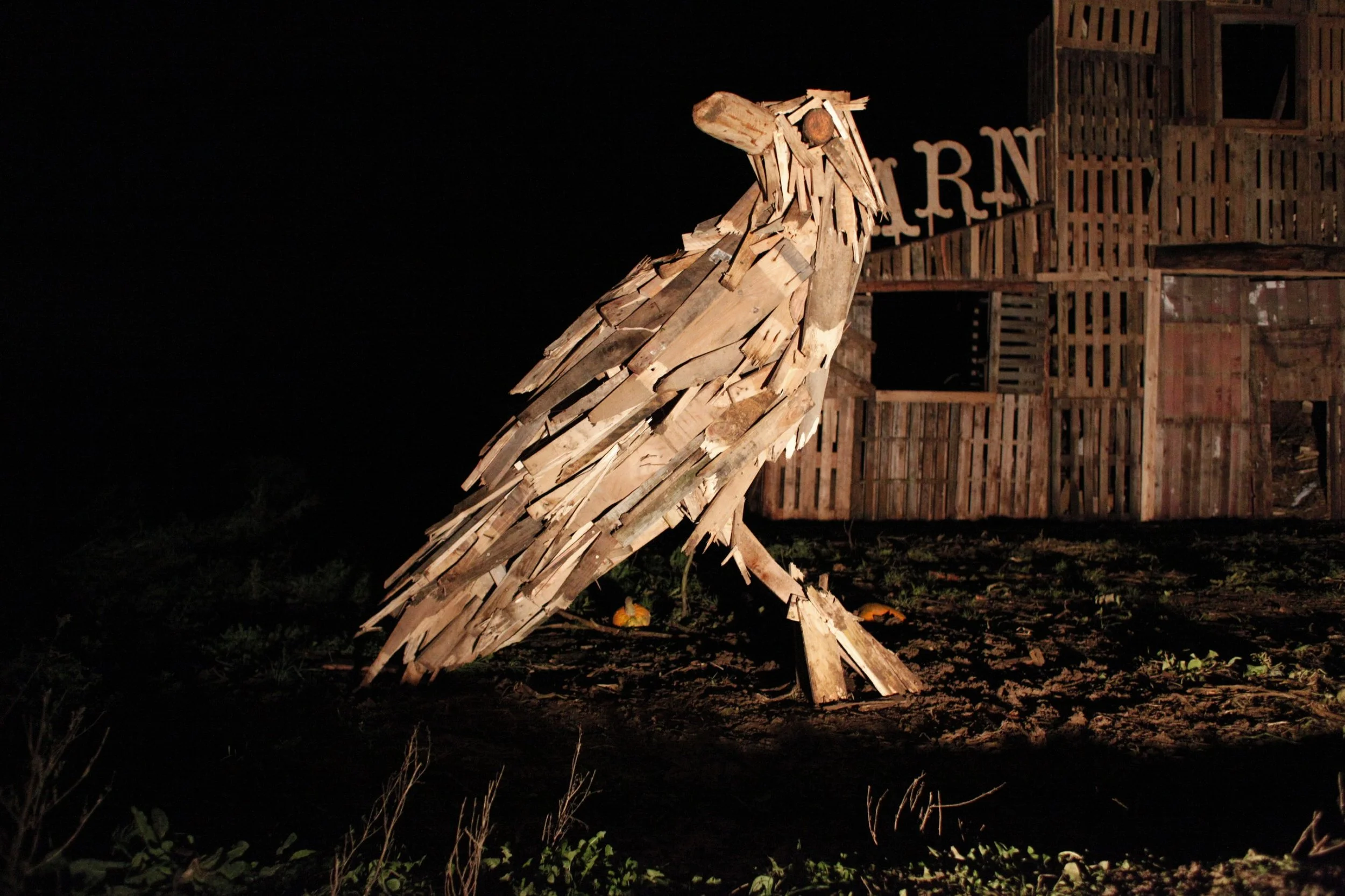 Wooden sculpture of a horse made from various wooden planks and pieces, situated outdoors at night with a wooden structure and illuminated signs in the background.